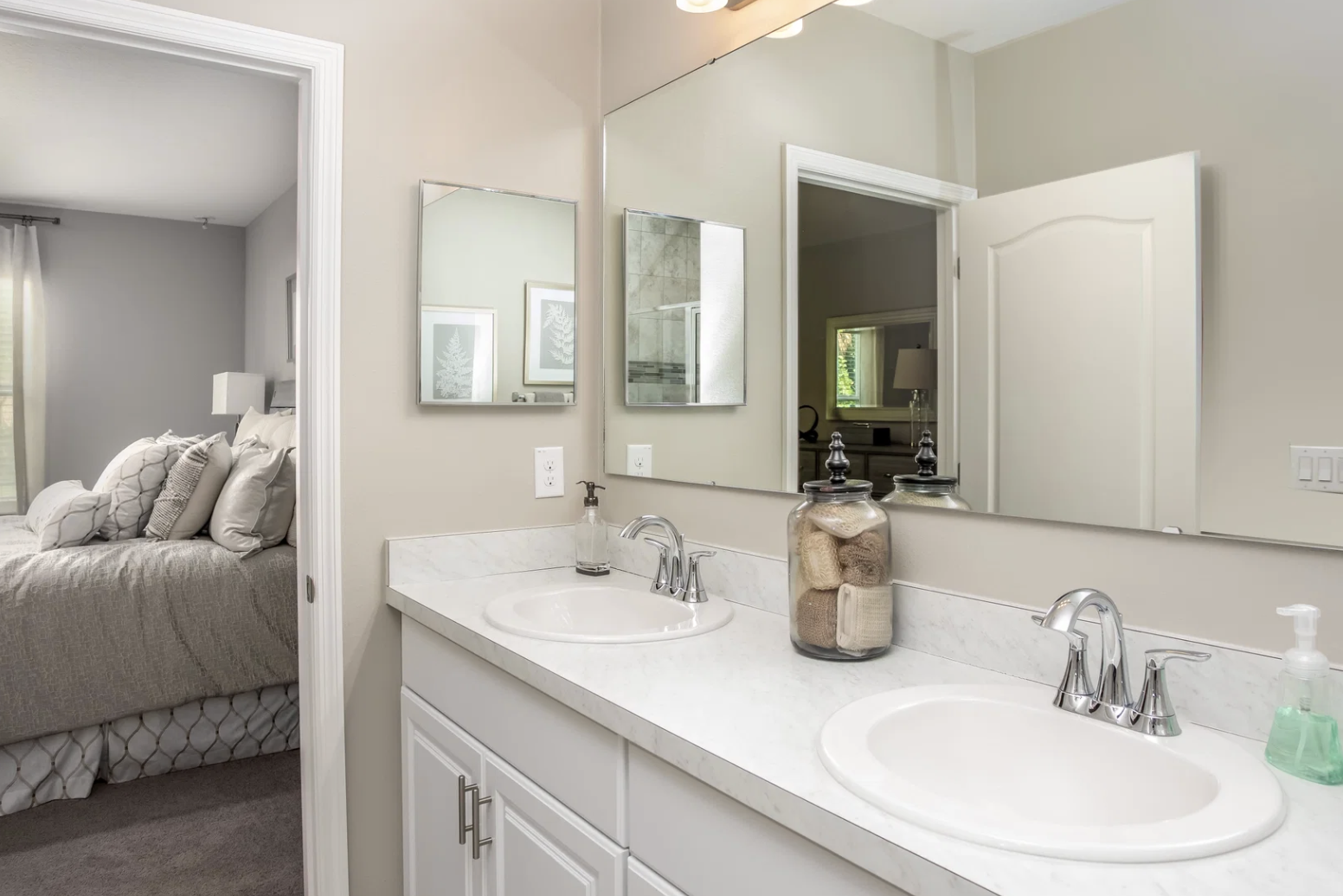 Spacious modern bathroom with double sinks, a mirror, and decorative storage jars, leading to a cozy bedroom.