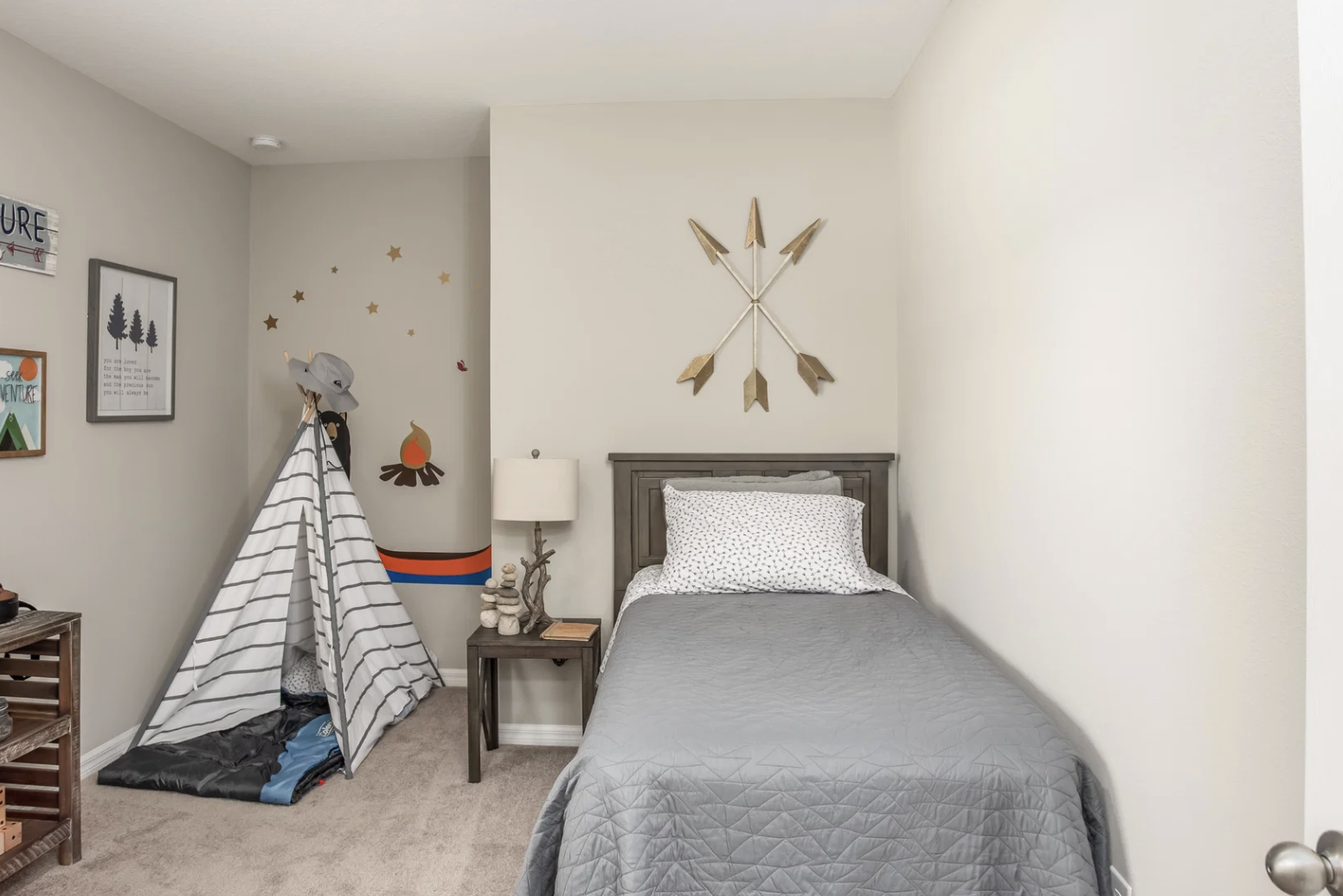Cozy children's bedroom featuring a gray bedspread, a decorative wall with arrows and stars, and a playful teepee in the corner.