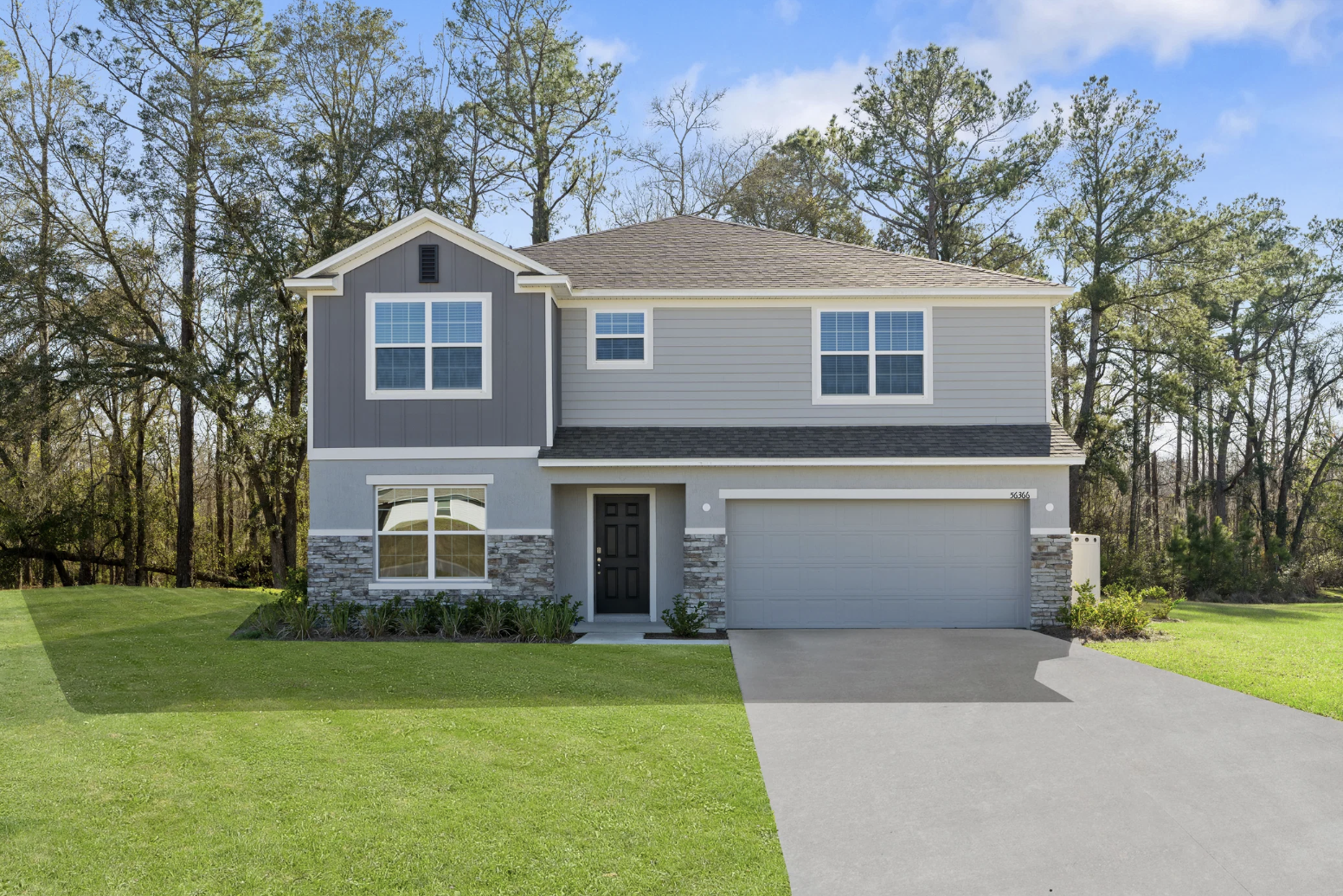 Two-story gray house with a stone accent, front lawn, and driveway, set against a backdrop of trees.