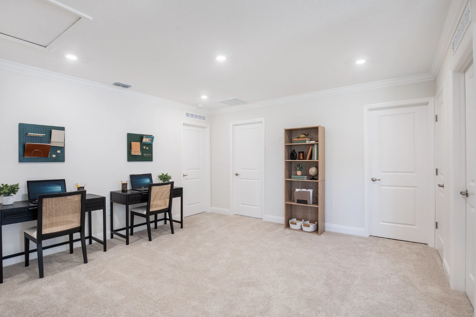 A softly lit room with two black desks, each with a chair, and a wooden bookshelf against the wall.