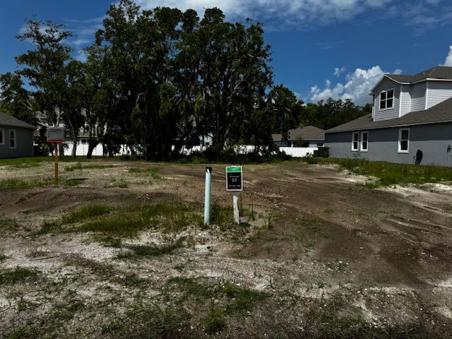 Image of a vacant residential lot with utility markers, surrounded by houses and trees, under a partly cloudy sky.