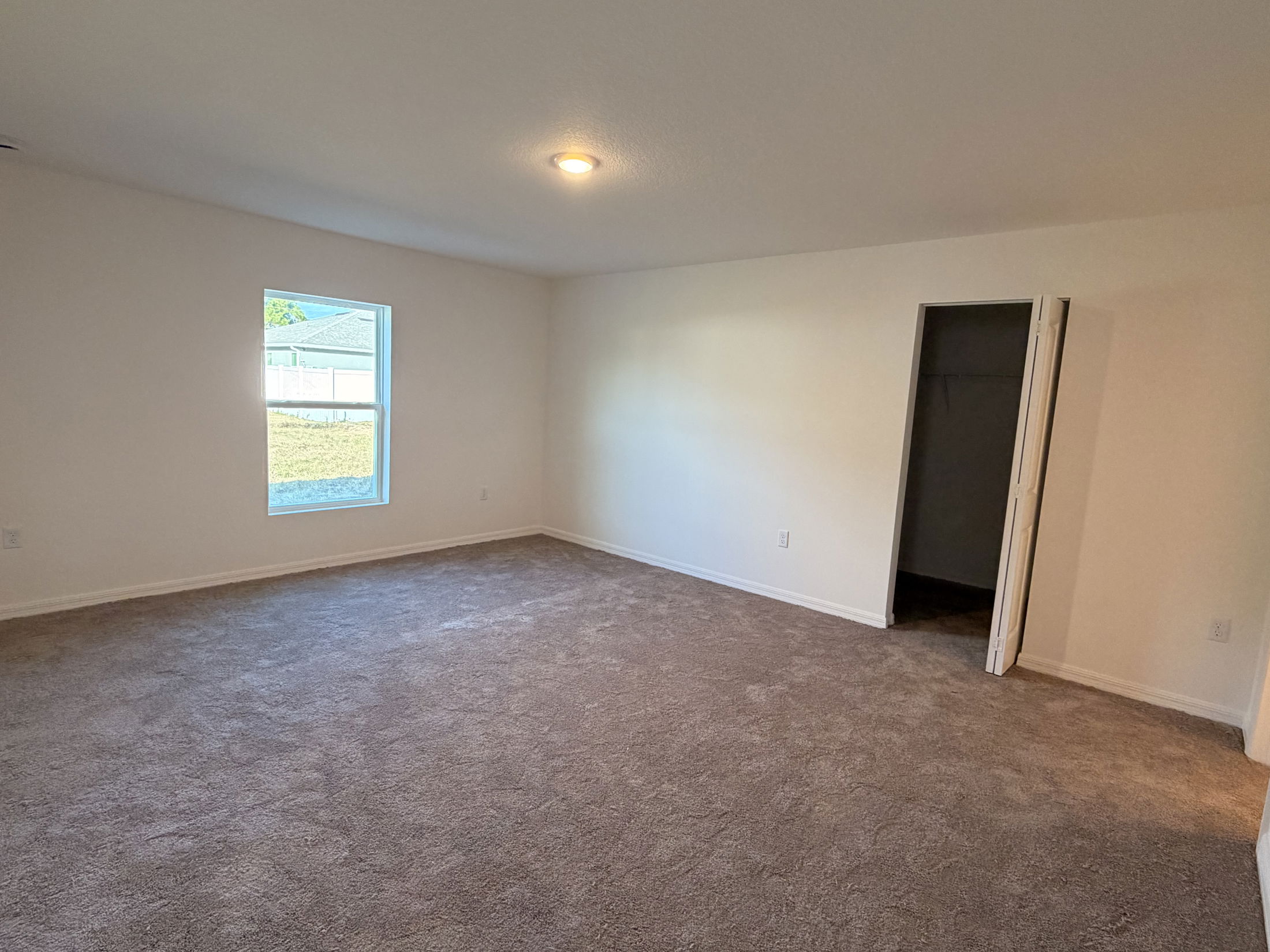 Spacious empty room with beige carpet, white walls, a large window, a ceiling light, and an open closet door.