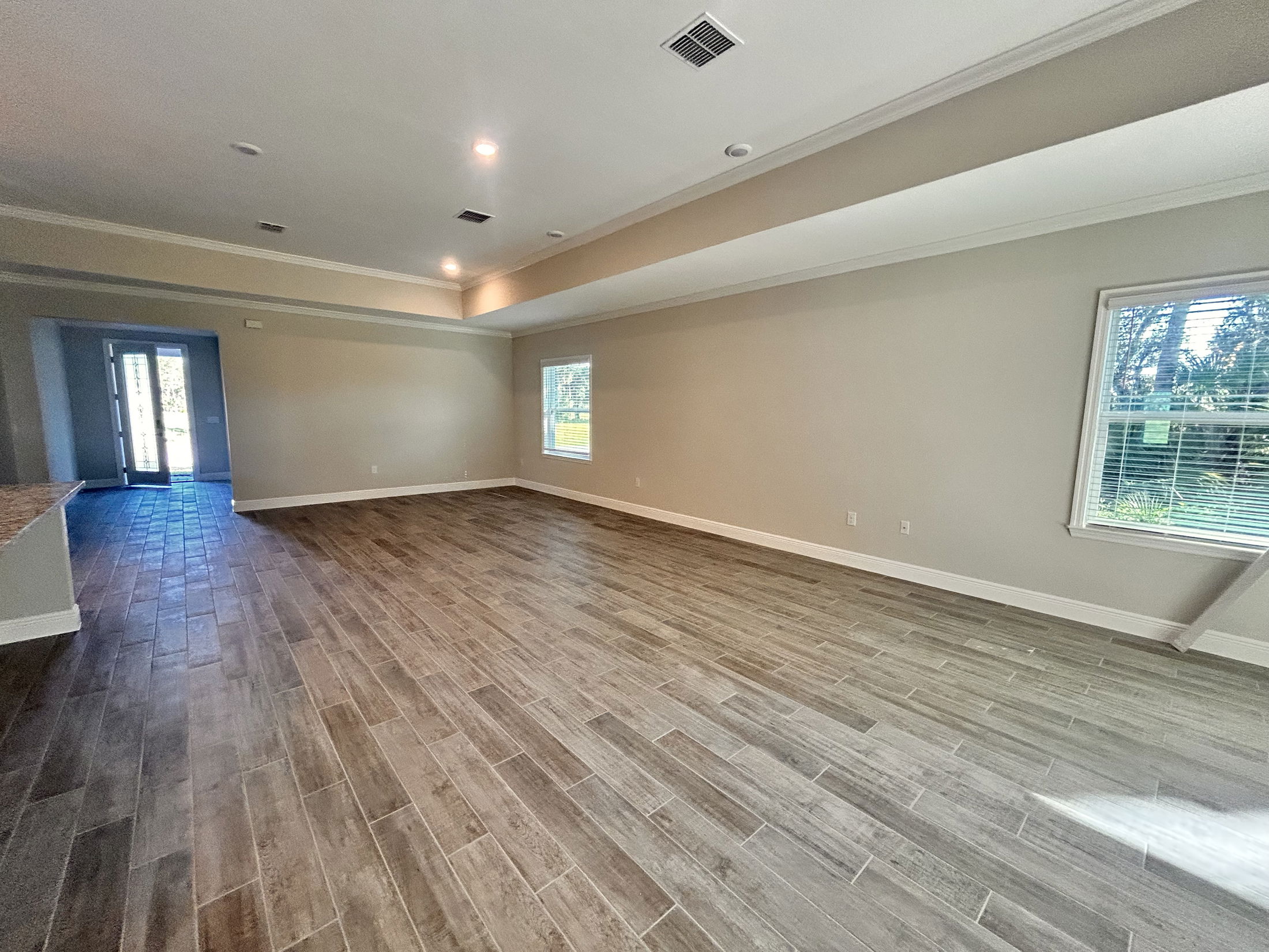 Spacious empty room with wood-like tile flooring, beige walls, and large windows letting in natural light.