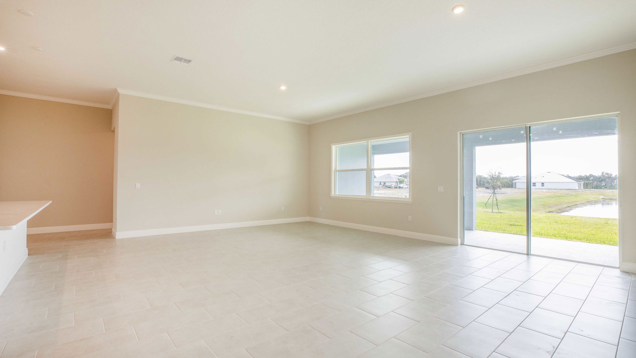 Spacious empty living room with beige walls, tile flooring, and large windows offering a view of a grassy yard.