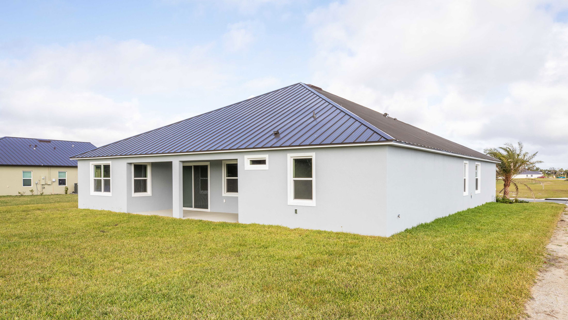 Modern single-story home with a blue metal roof and light blue exterior, surrounded by a green lawn under a cloudy sky.
