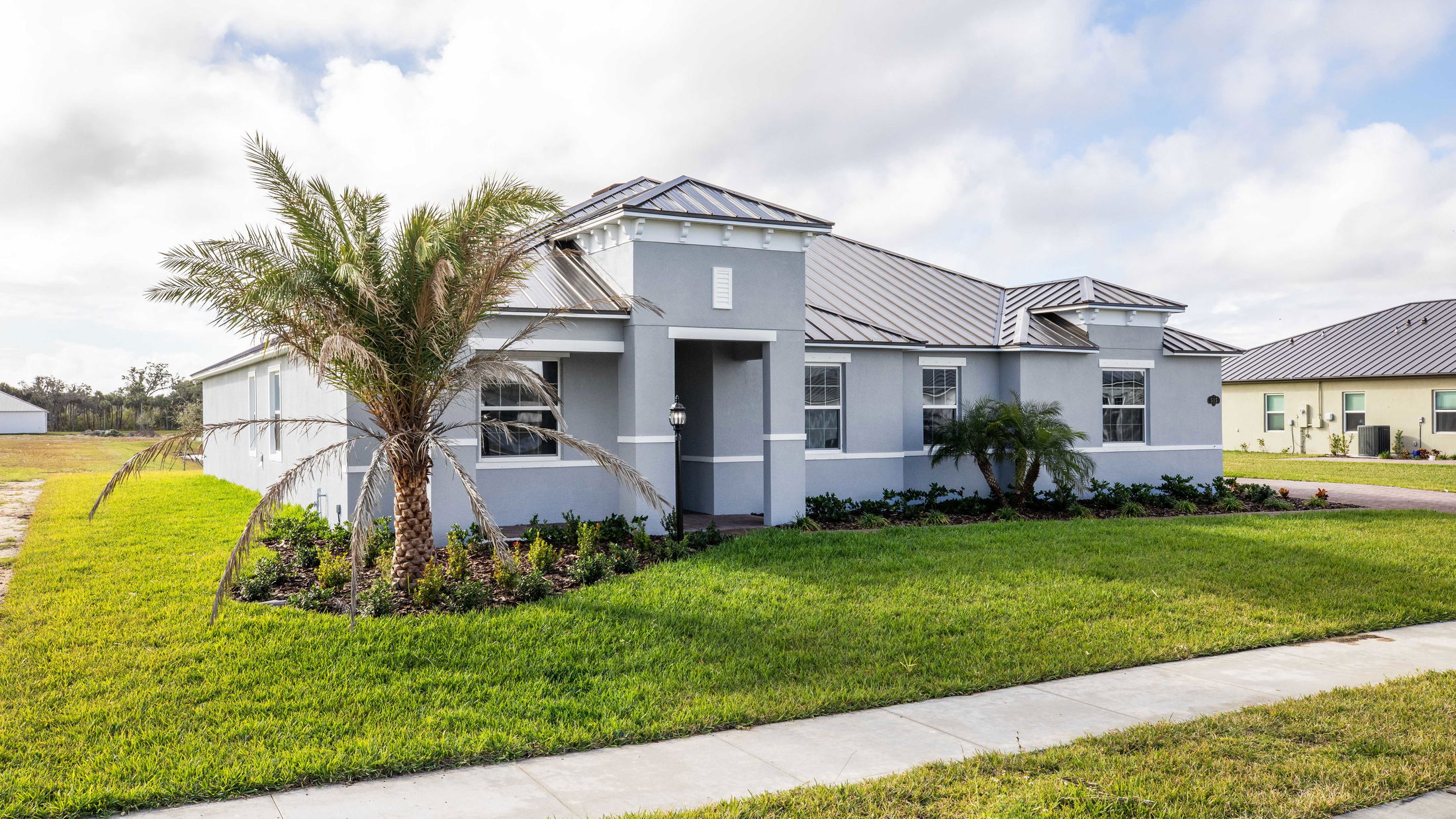 Modern single-story gray house with a metal roof and lush green lawn featuring palm trees and shrubs in the front yard against a partly cloudy sky.
