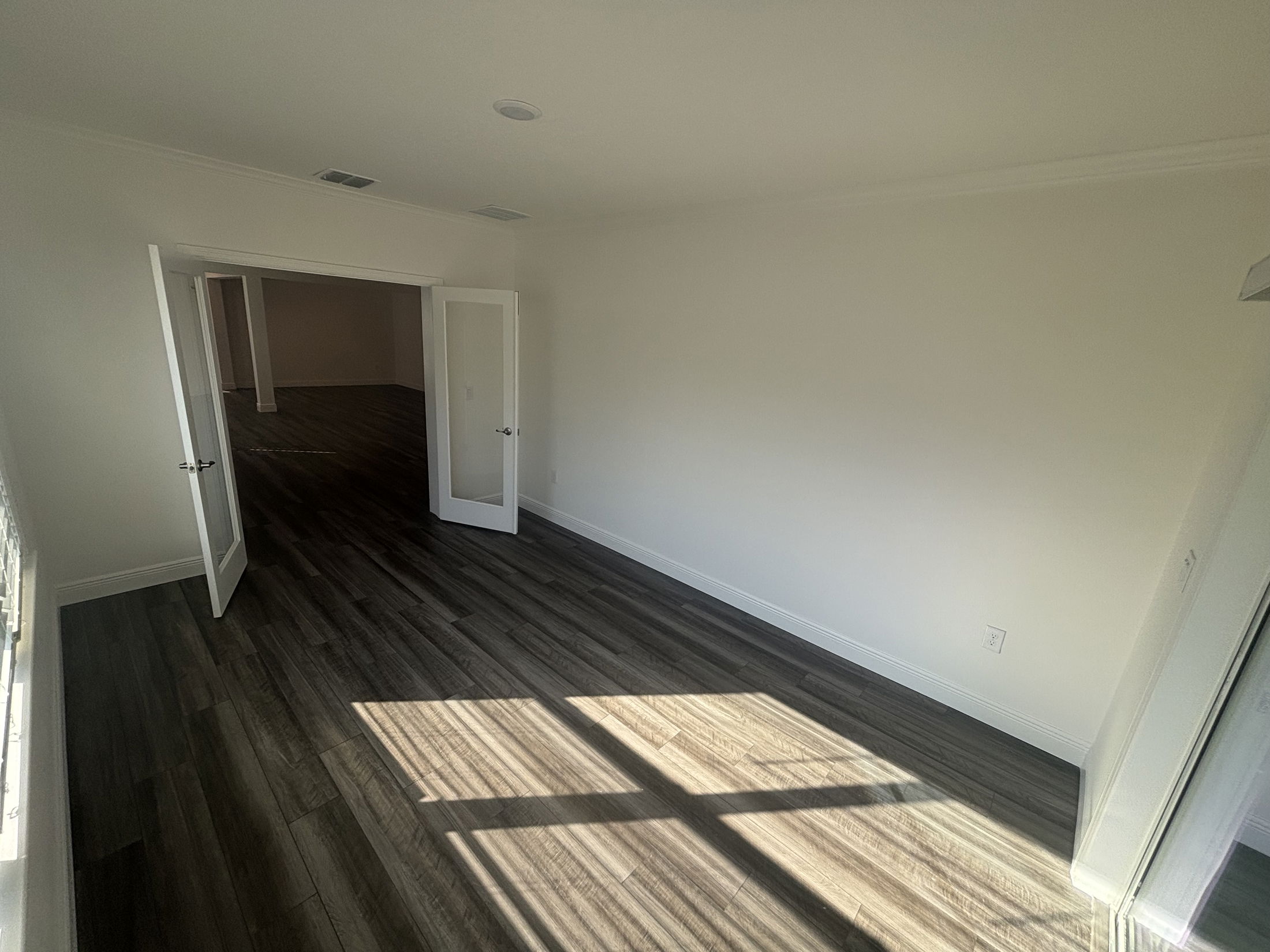 Bright, empty room with dark wood flooring, white walls, and French doors opening to another space.