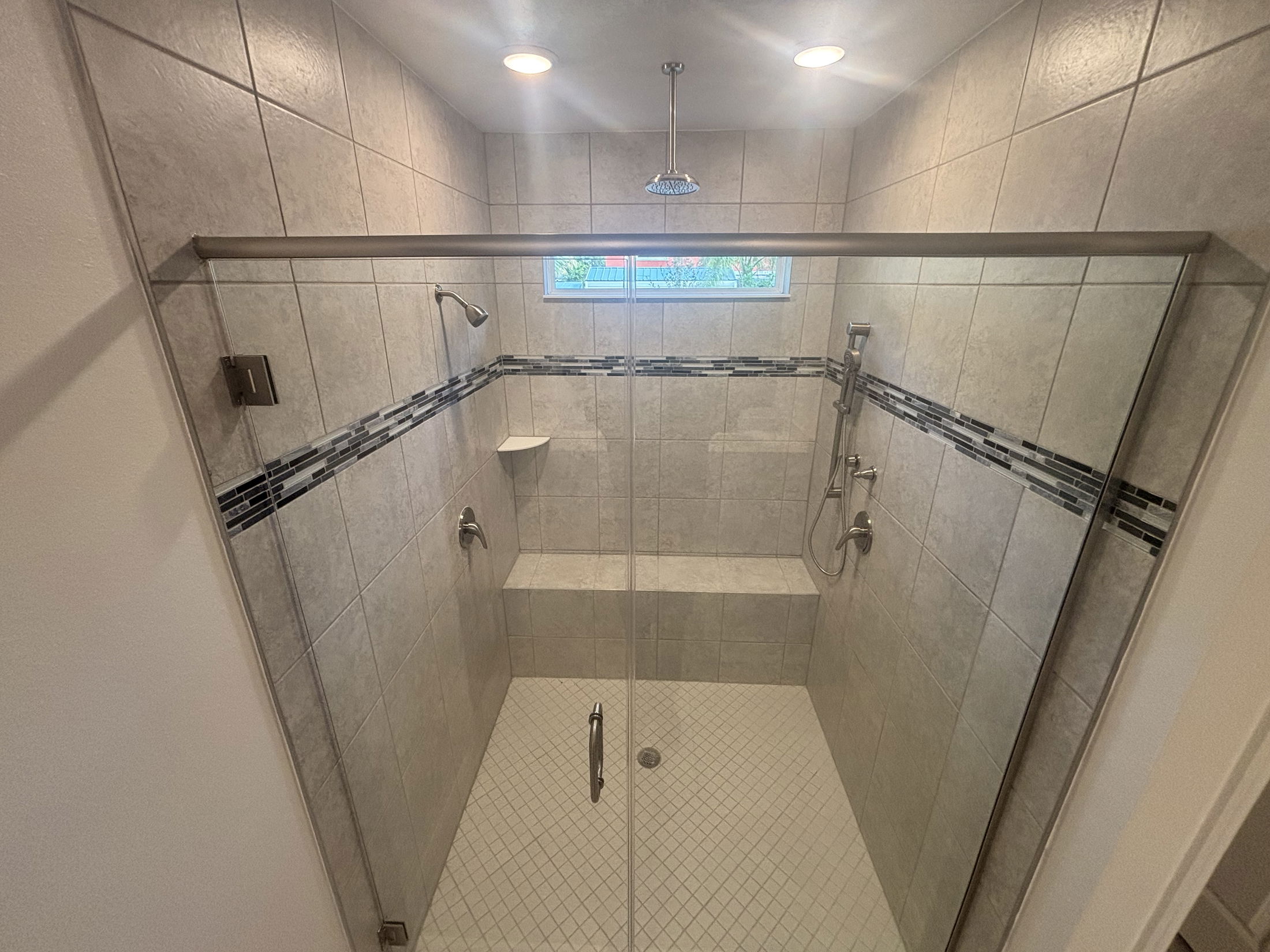 Elegant modern tiled shower with glass door featuring a rainfall showerhead and convenient built-in bench.