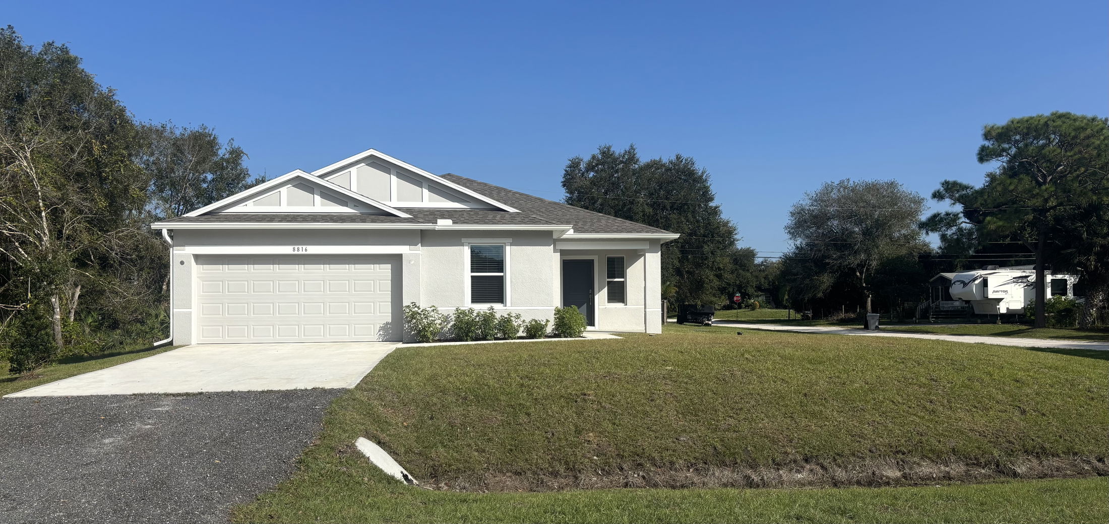 Single-story white house with a two-car garage, surrounded by trees, under a clear blue sky.