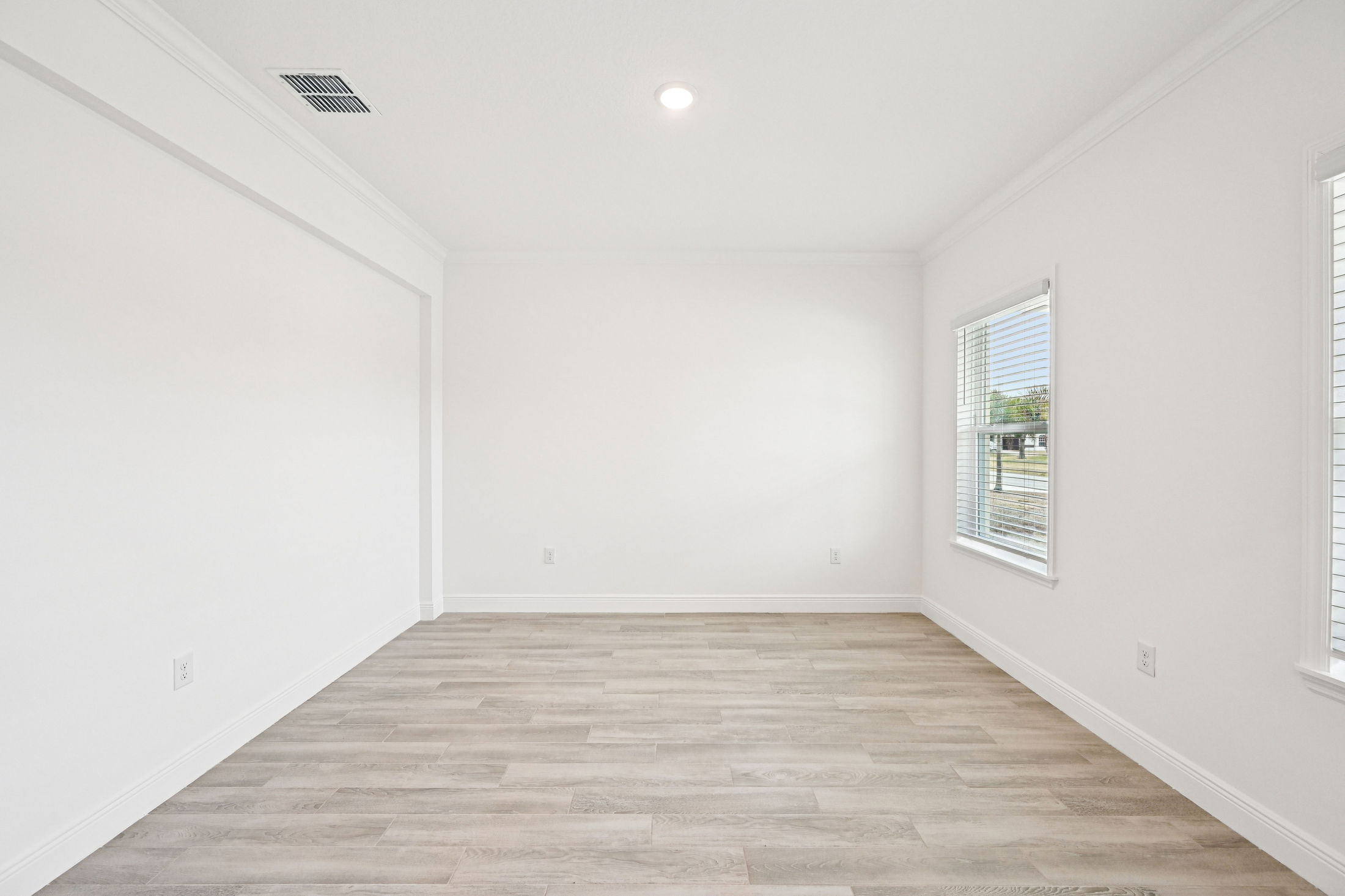 Spacious and bright empty room with white walls and wood-like flooring, featuring windows with blinds.