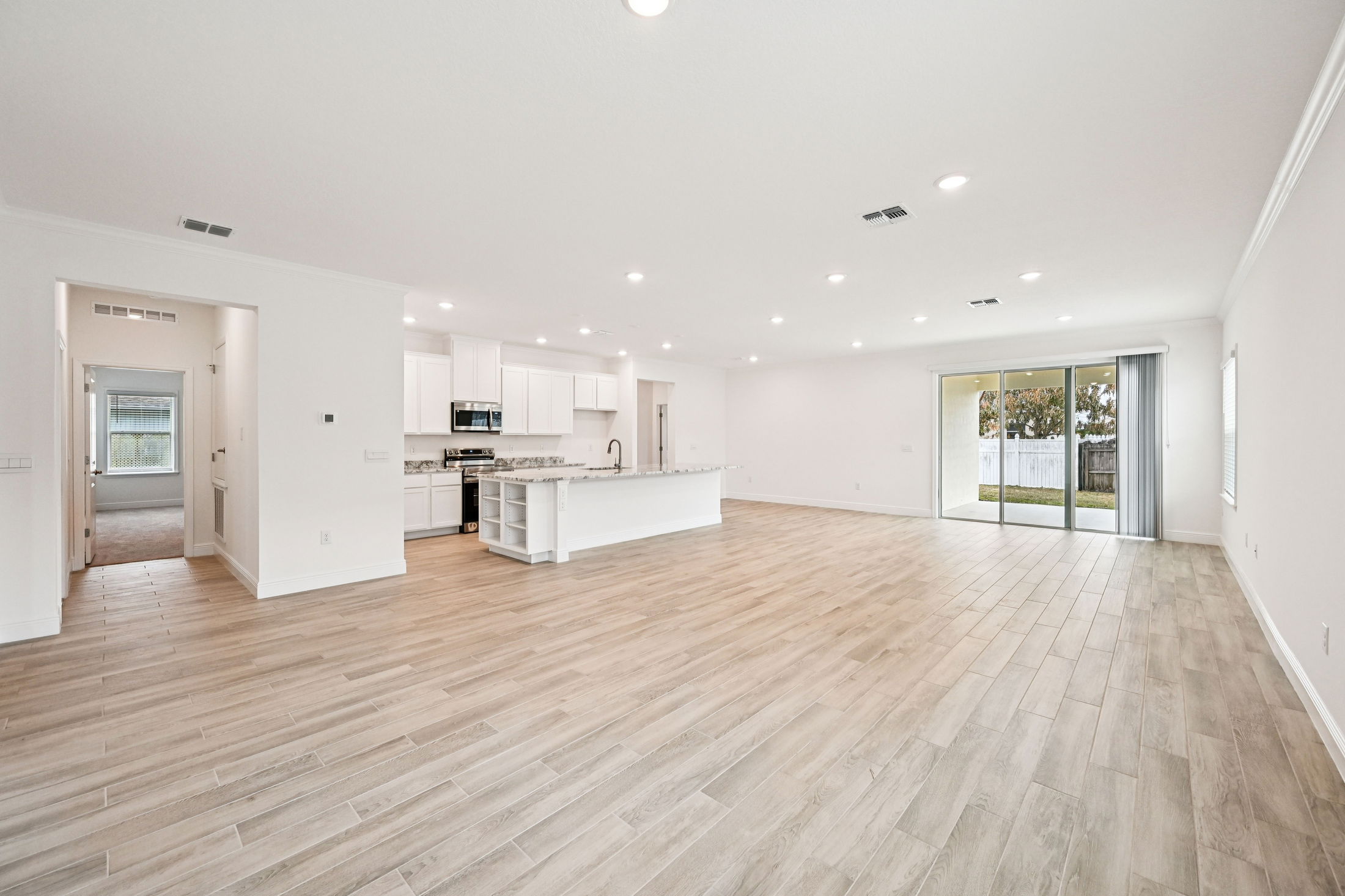 Spacious modern living room with open kitchen layout featuring light wood flooring and large sliding glass doors to the backyard.