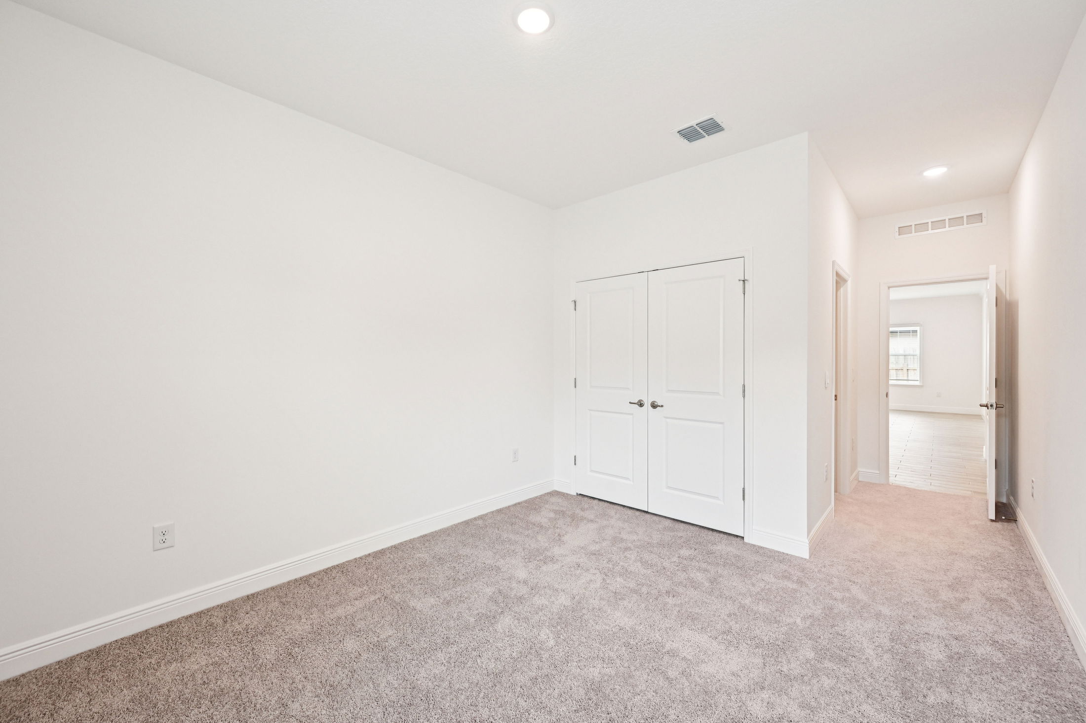 Spacious and bright empty bedroom with beige carpet and white walls featuring double doors and a doorway leading to another room.