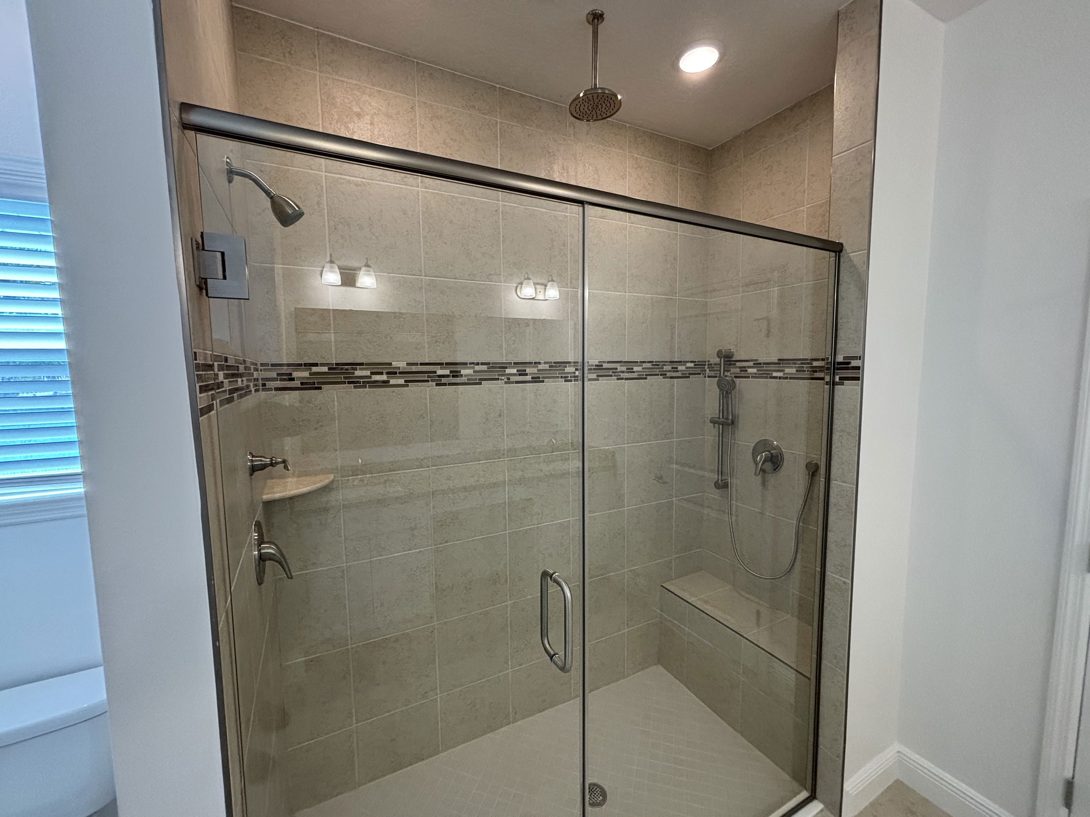 Modern glass-enclosed shower with dual shower heads and beige tiled walls featuring decorative accent tiles.