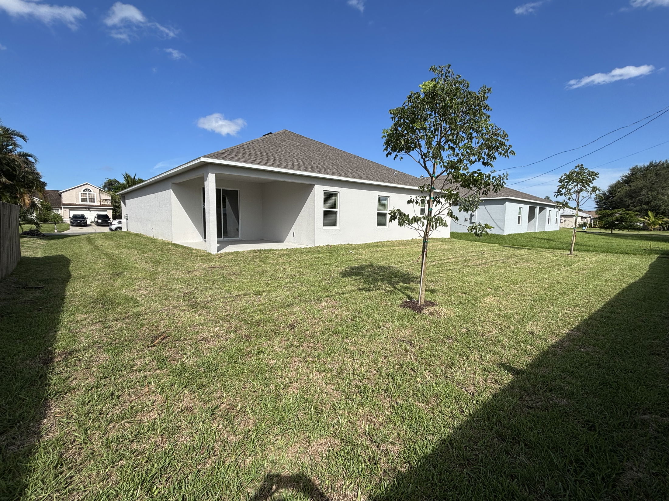 Backyard of a suburban house with a large grassy lawn, two young trees, and a covered patio under a clear blue sky.