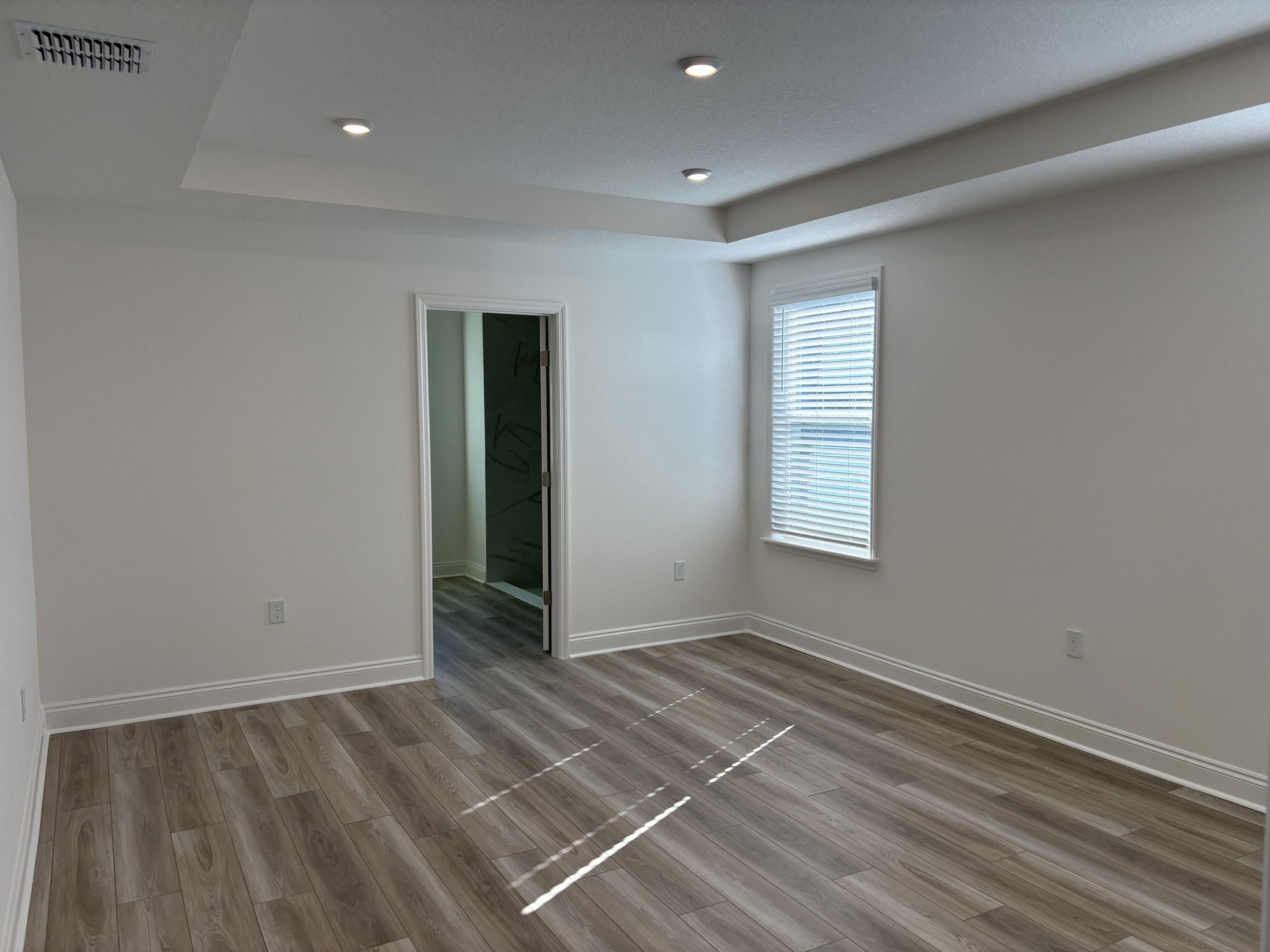 Spacious and bright empty room with wooden flooring, a window with blinds, and a doorway leading to another space.