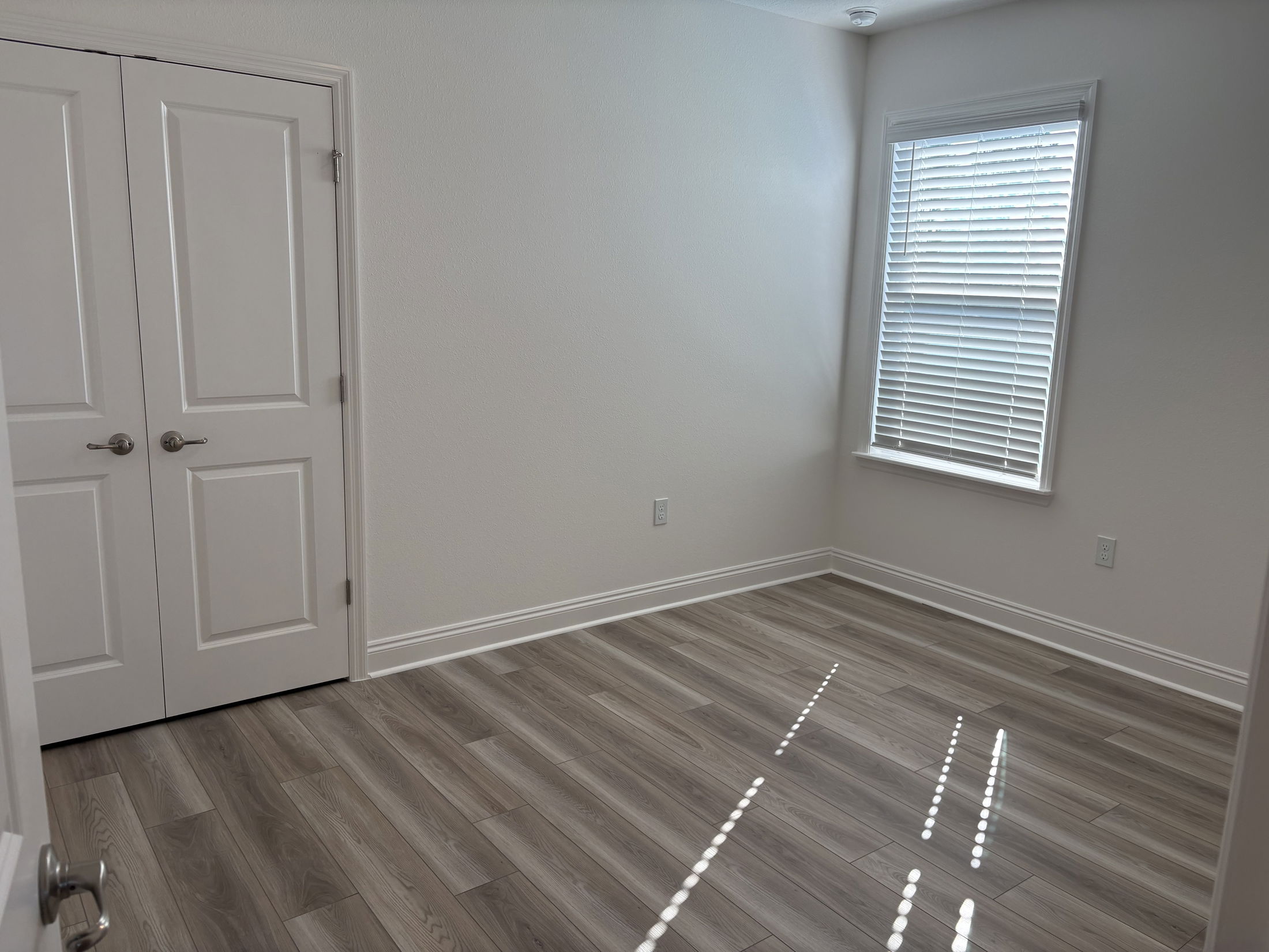 Modern empty room with natural light, gray wood flooring, and white walls featuring double doors and window blinds.