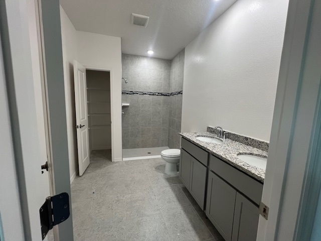 Modern bathroom with dual-sink granite countertop, gray cabinets, tiled shower, and open closet.