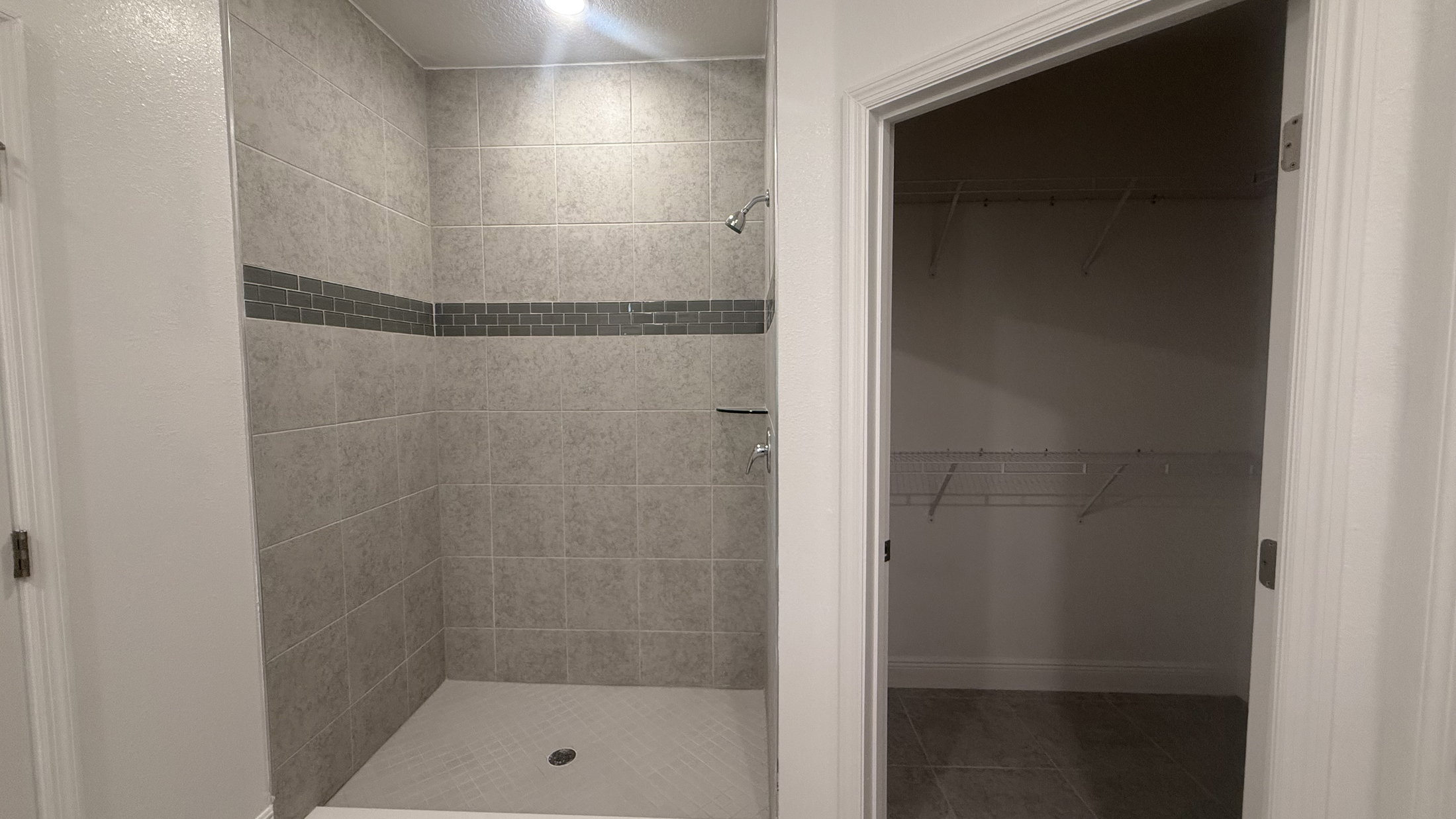 A modern shower stall with gray tiled walls and a glass door, adjacent to an empty closet with shelves.