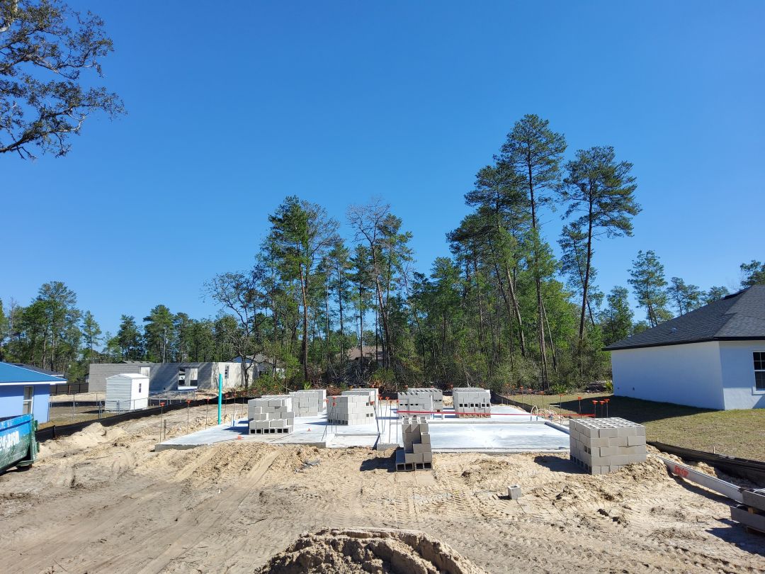 Construction site with foundation and building materials surrounded by trees under a clear blue sky.