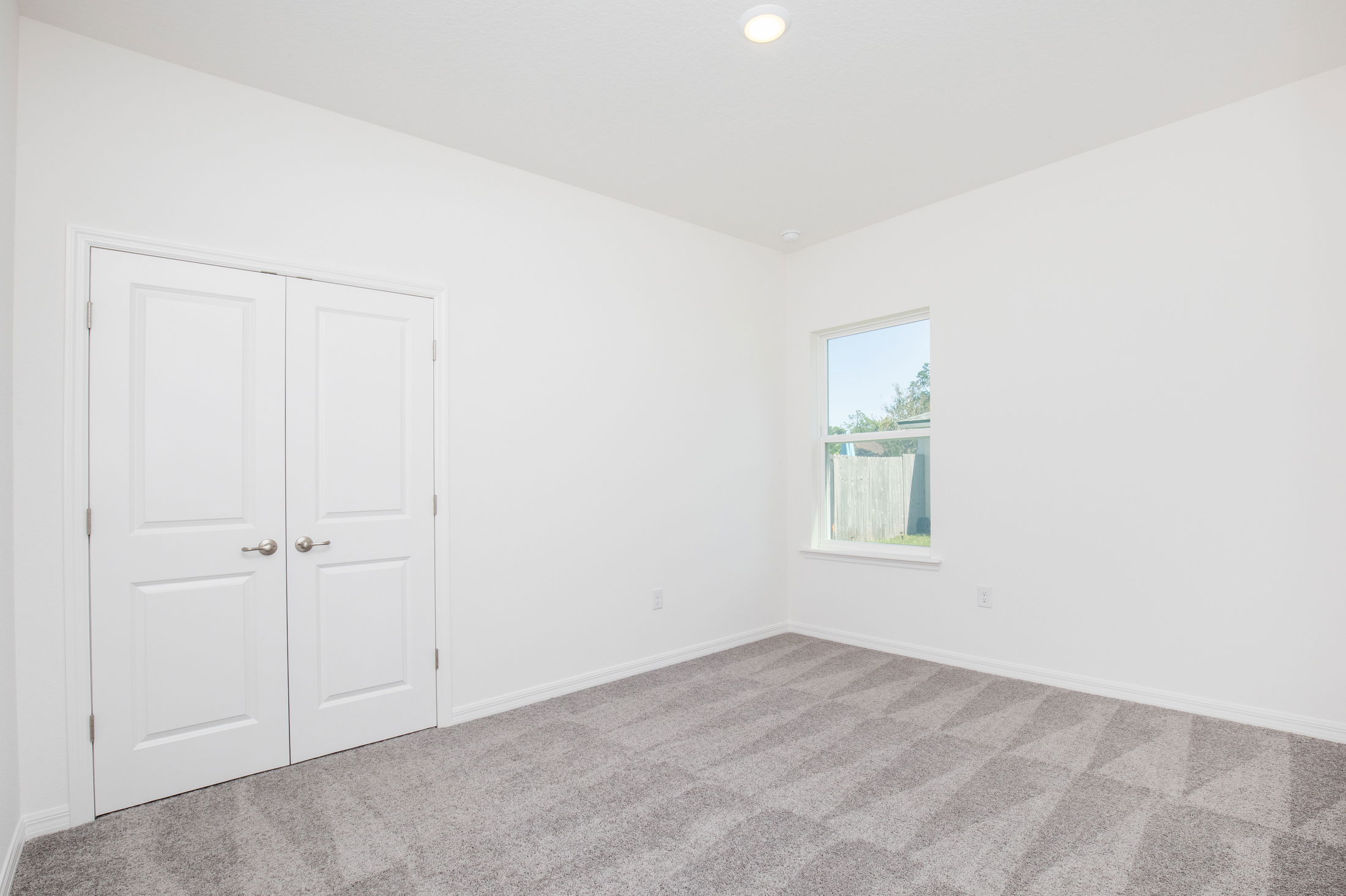 Empty white bedroom with carpet flooring, double doors, and a window overlooking a backyard.