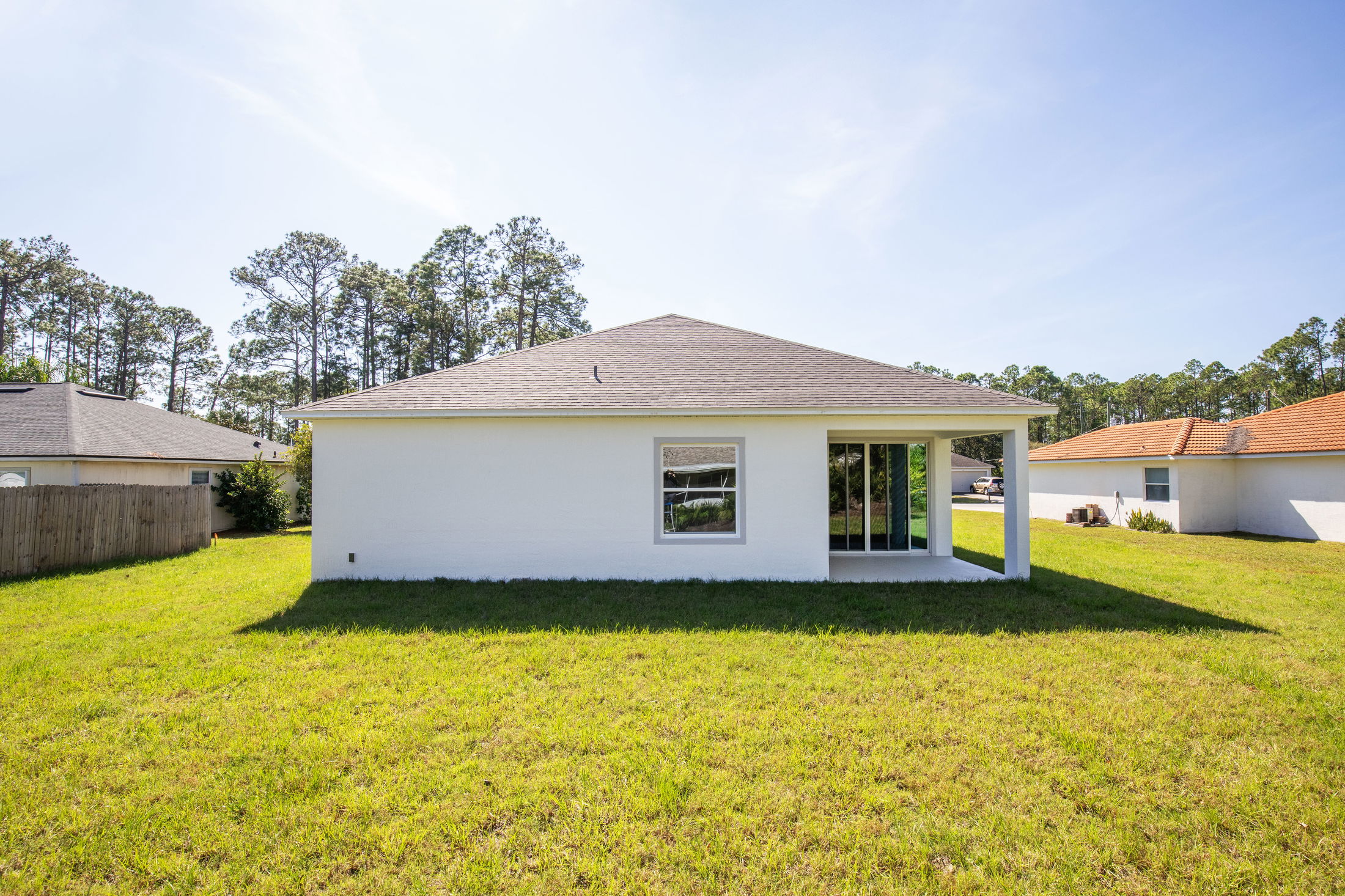 Back view of a single-story white house with a patio and a grassy backyard, surrounded by a suburban neighborhood with trees.
