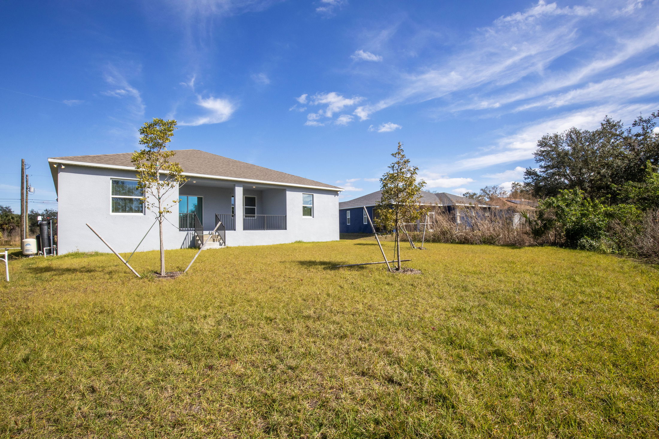 Suburban backyard with grassy lawn, young trees, and a modern single-story house under a clear blue sky.