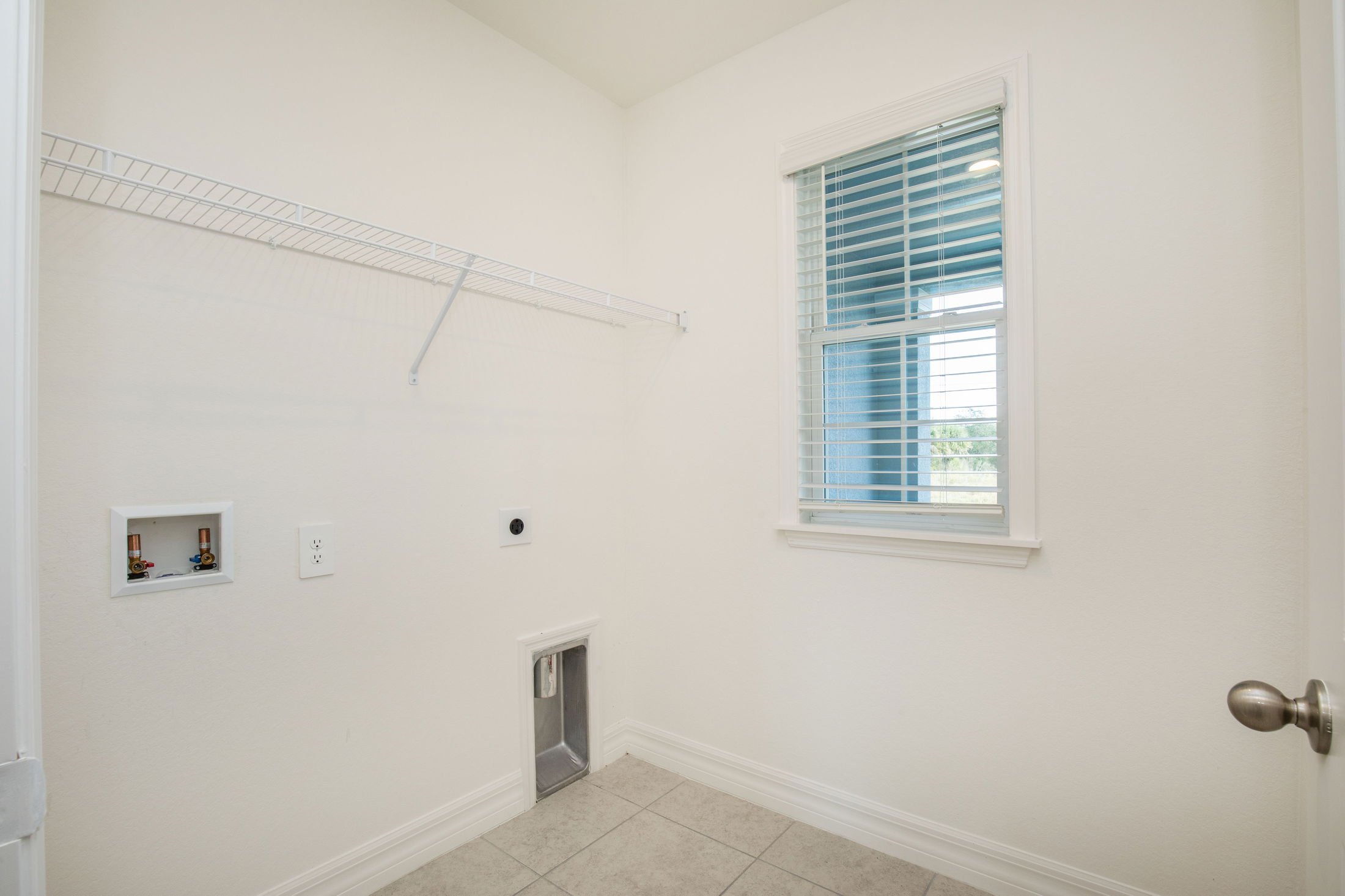 Empty laundry room with white walls, tiled floor, window with blinds, and a wire shelving unit.