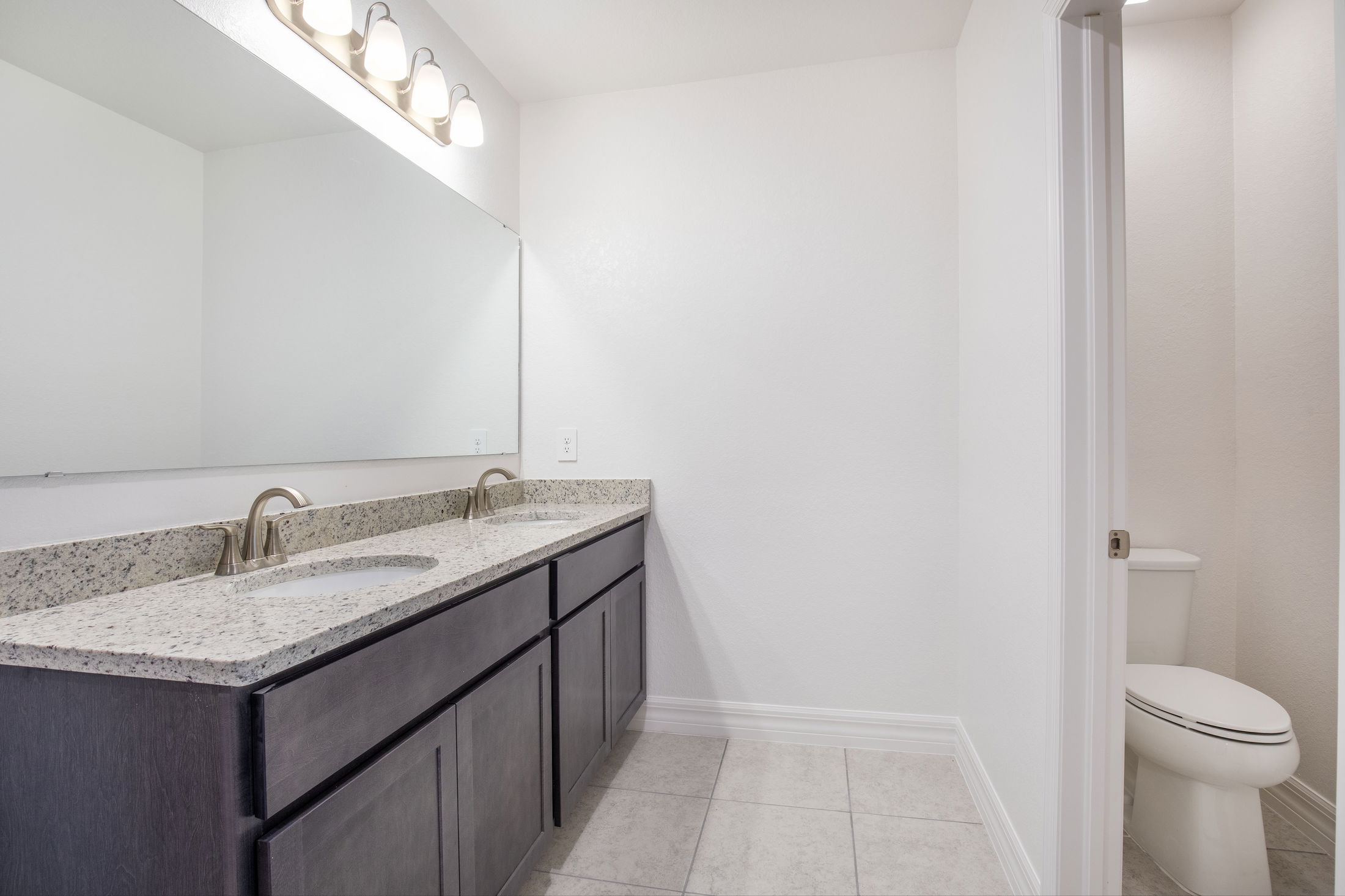 Modern bathroom with dual sinks, granite countertop, large mirror, and adjacent toilet in a bright, tiled space.