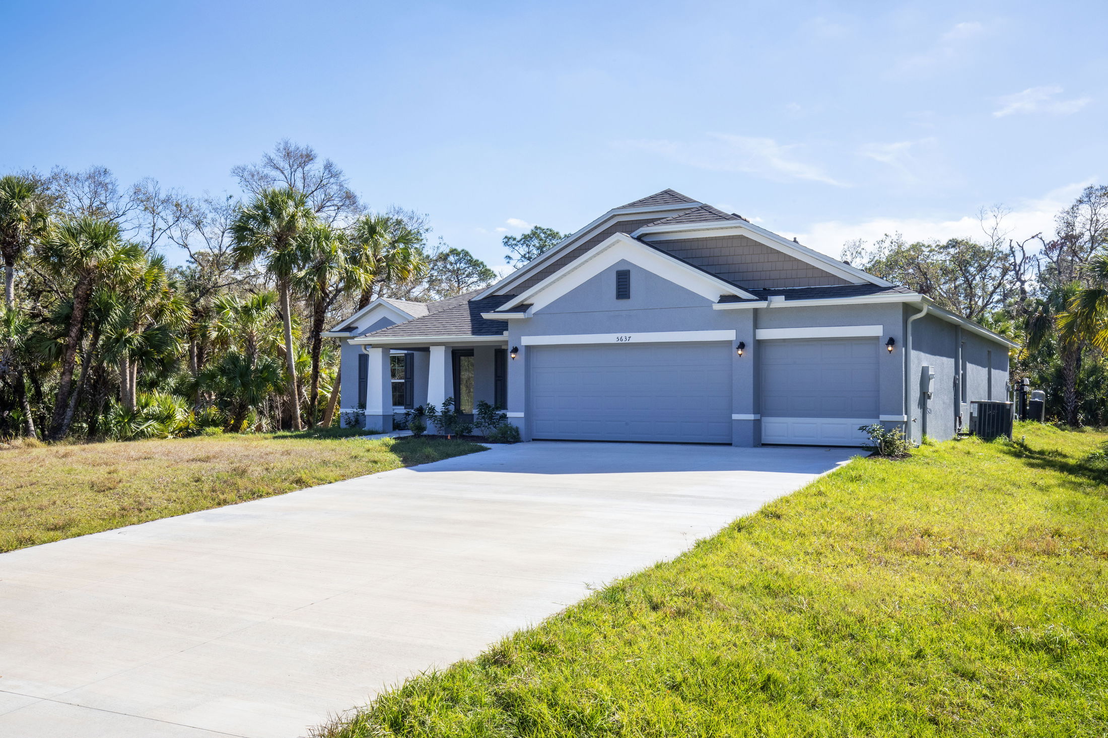 Modern gray suburban home with a spacious driveway, surrounded by lush greenery and palm trees.