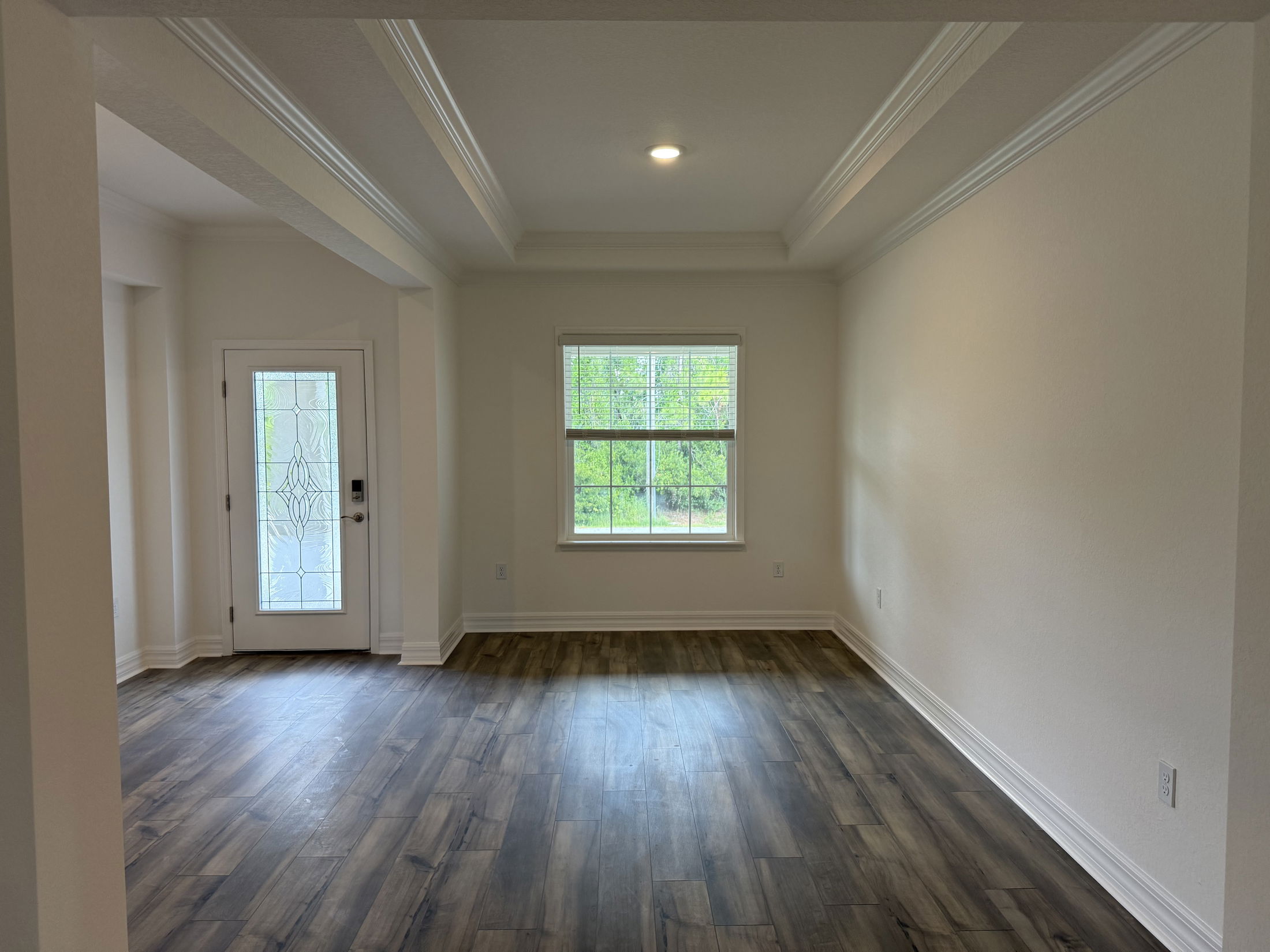 Empty room with wooden flooring, white walls, and a decorative glass front door adjacent to a large window.