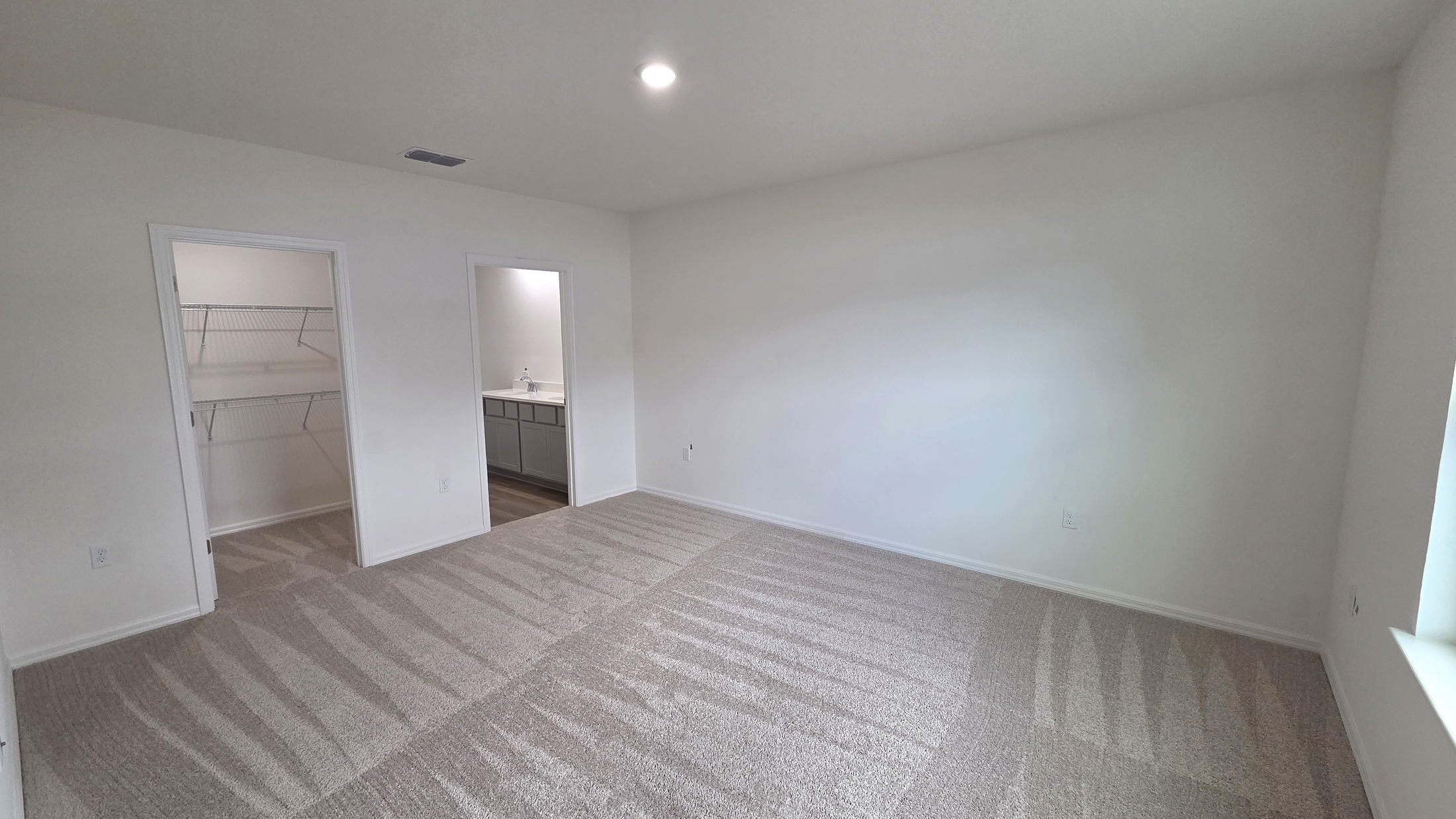 Spacious and well-lit bedroom featuring neutral walls, plush carpeting, and adjoining closets with a view of a modern bathroom.