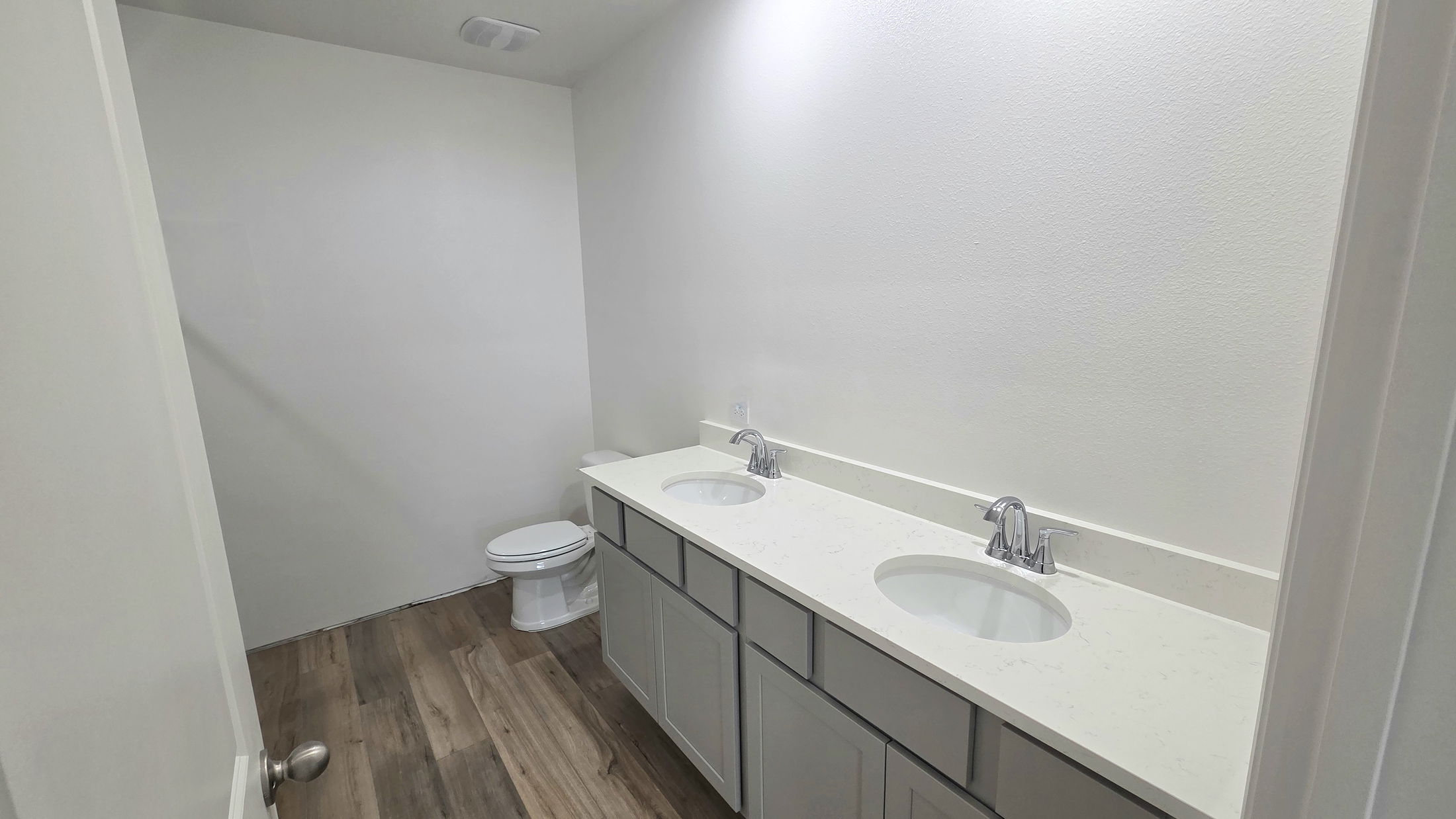Modern bathroom design featuring dual sinks, a toilet, and a simple, clean aesthetic with light-colored cabinetry and wood-look flooring.