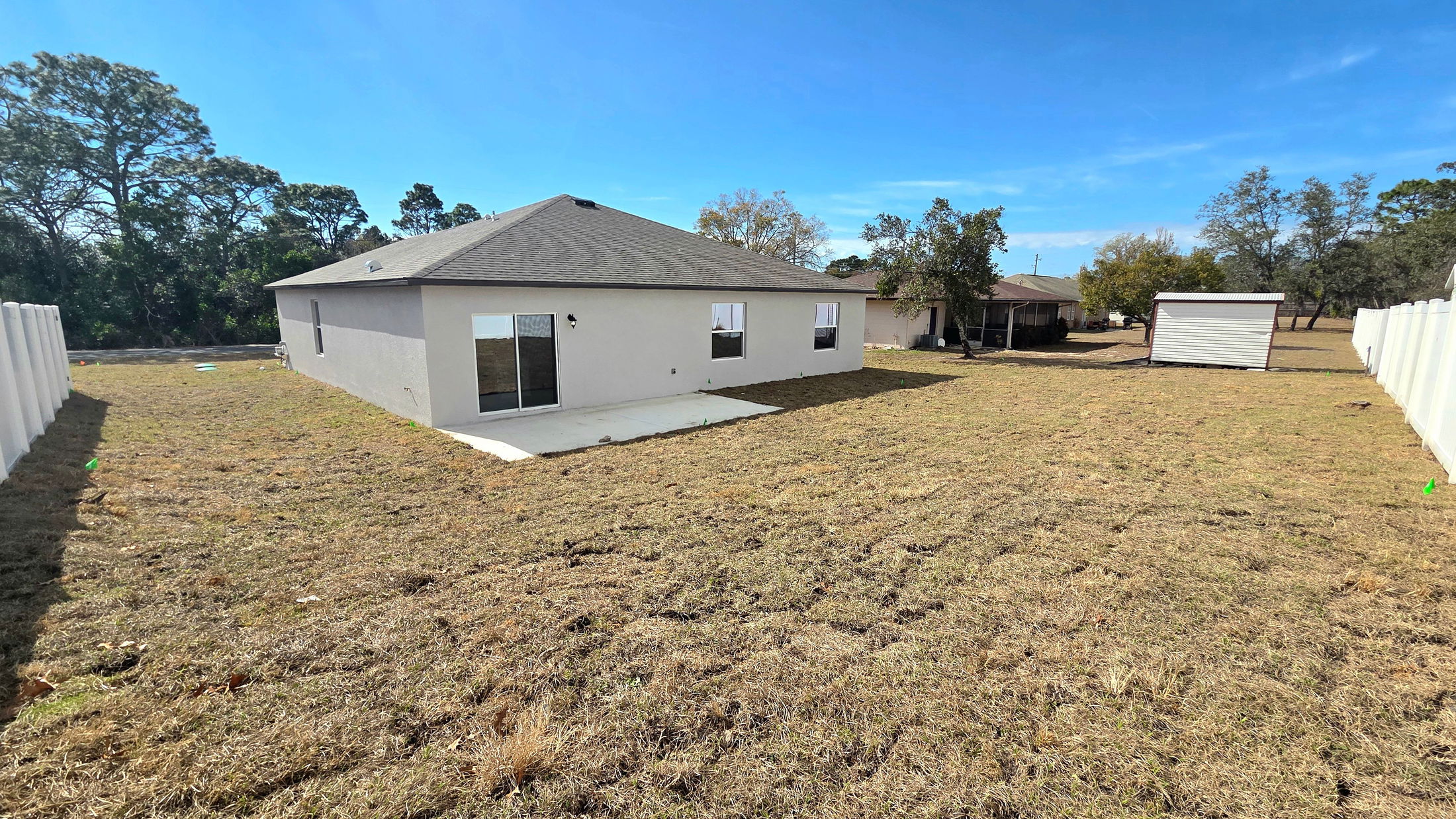 A spacious backyard view of a modern home featuring a concrete patio and a storage shed against a clear blue sky.