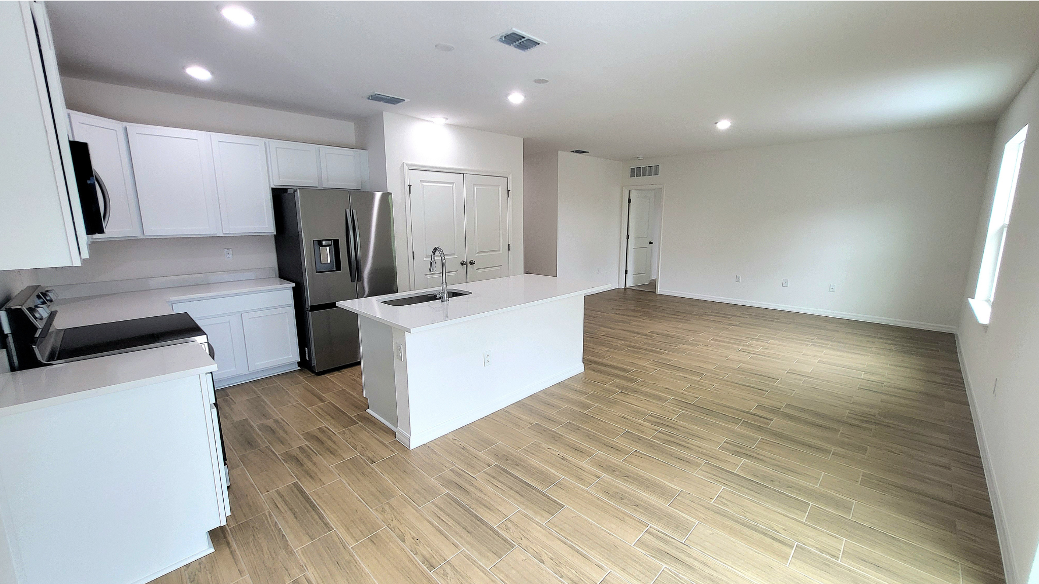 Modern open-concept kitchen with white cabinets, stainless steel appliances, and a spacious living area featuring light wood tile flooring.