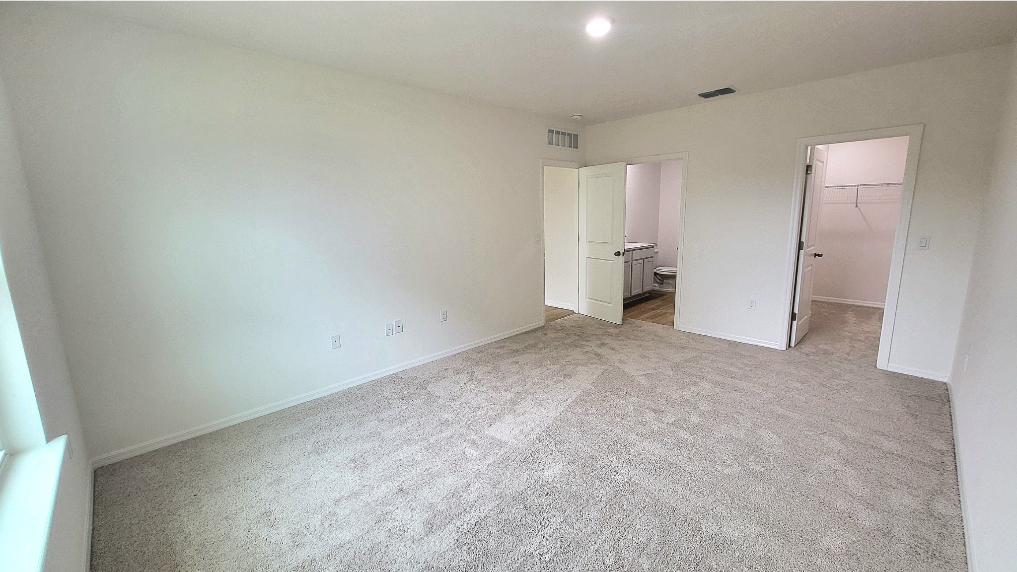 Bright, empty room with beige carpet, white walls, and open doors leading to a bathroom and closet.