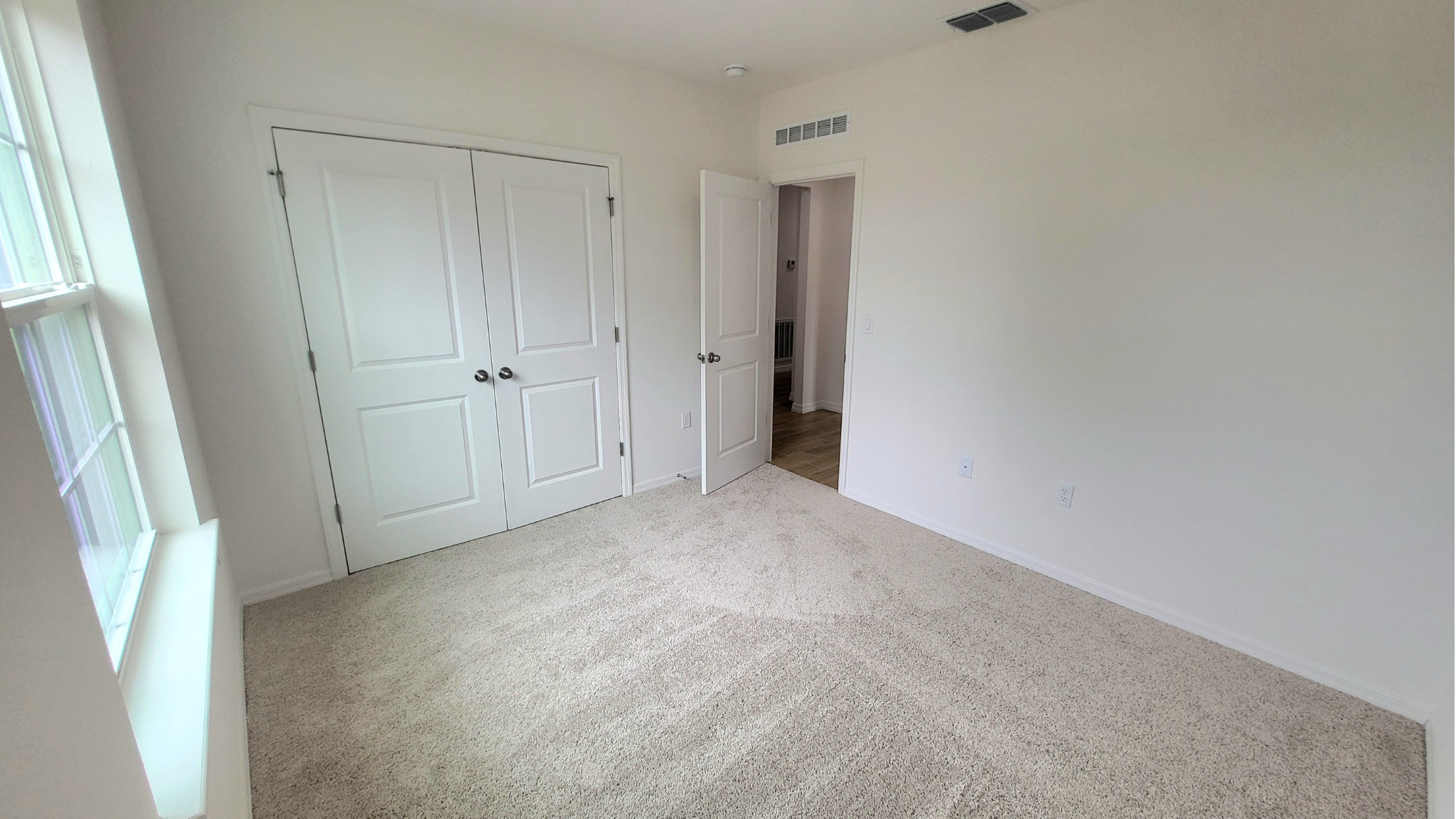 Empty bedroom with beige carpet, white walls, a double-door closet, a window, and an open door leading to a hallway.