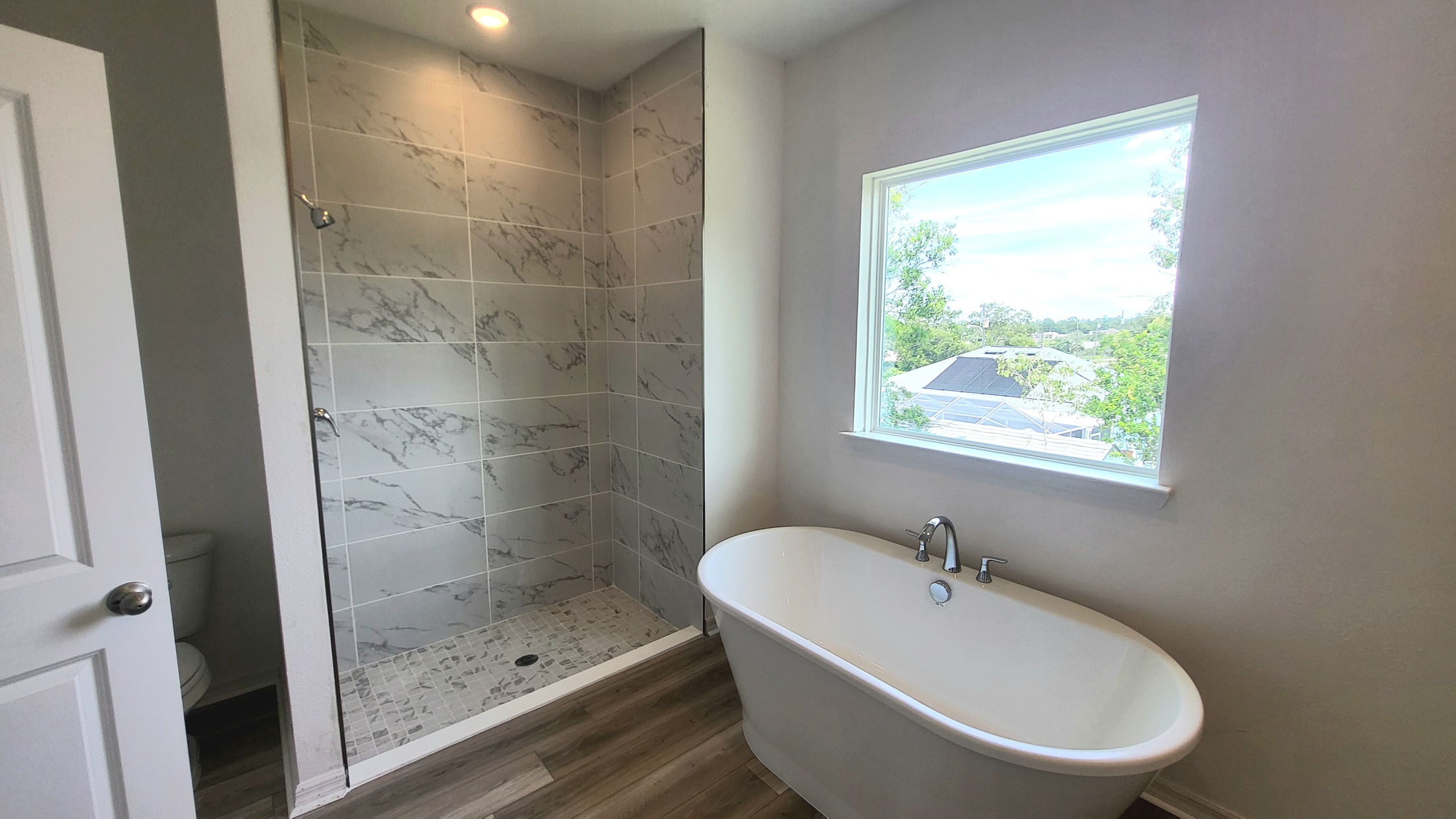 Modern bathroom featuring a luxurious freestanding bathtub, spacious walk-in shower with gray marble tiles, and a large window with an outdoor view.