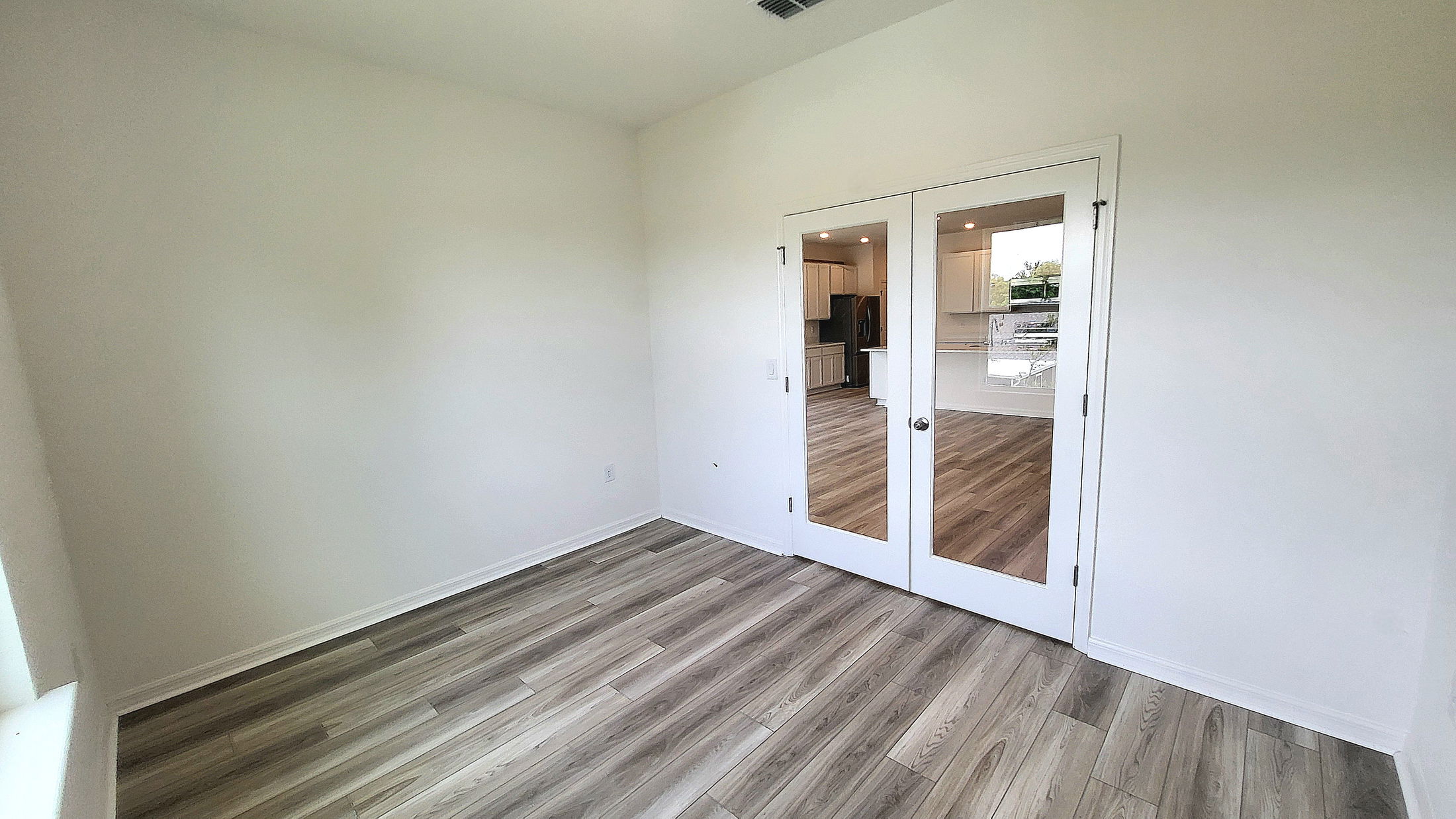 Empty room with light wood flooring and white walls, featuring glass French doors leading to a bright kitchen area.