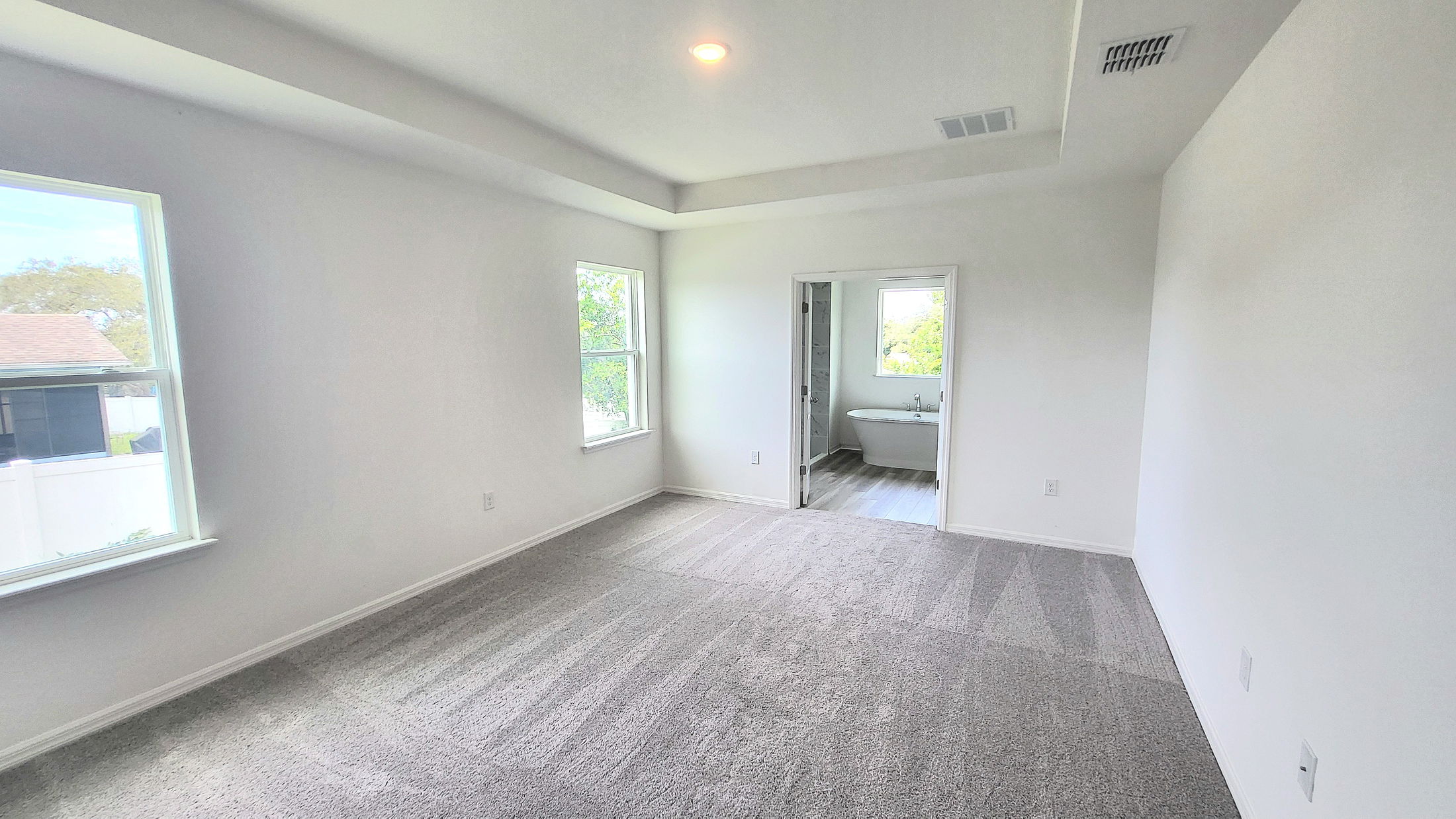 Spacious, empty bedroom with carpet flooring, large windows, and an en-suite bathroom featuring a modern bathtub.