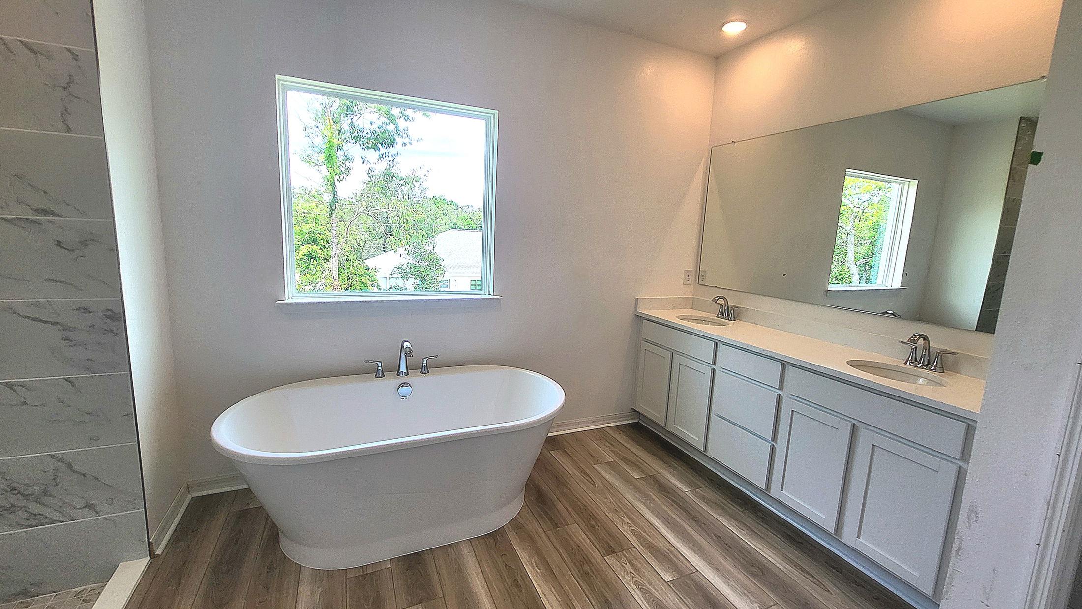 Modern bathroom with a standalone bathtub, large mirror, and dual-sink vanity, featuring wood-look flooring and a window with a natural view.