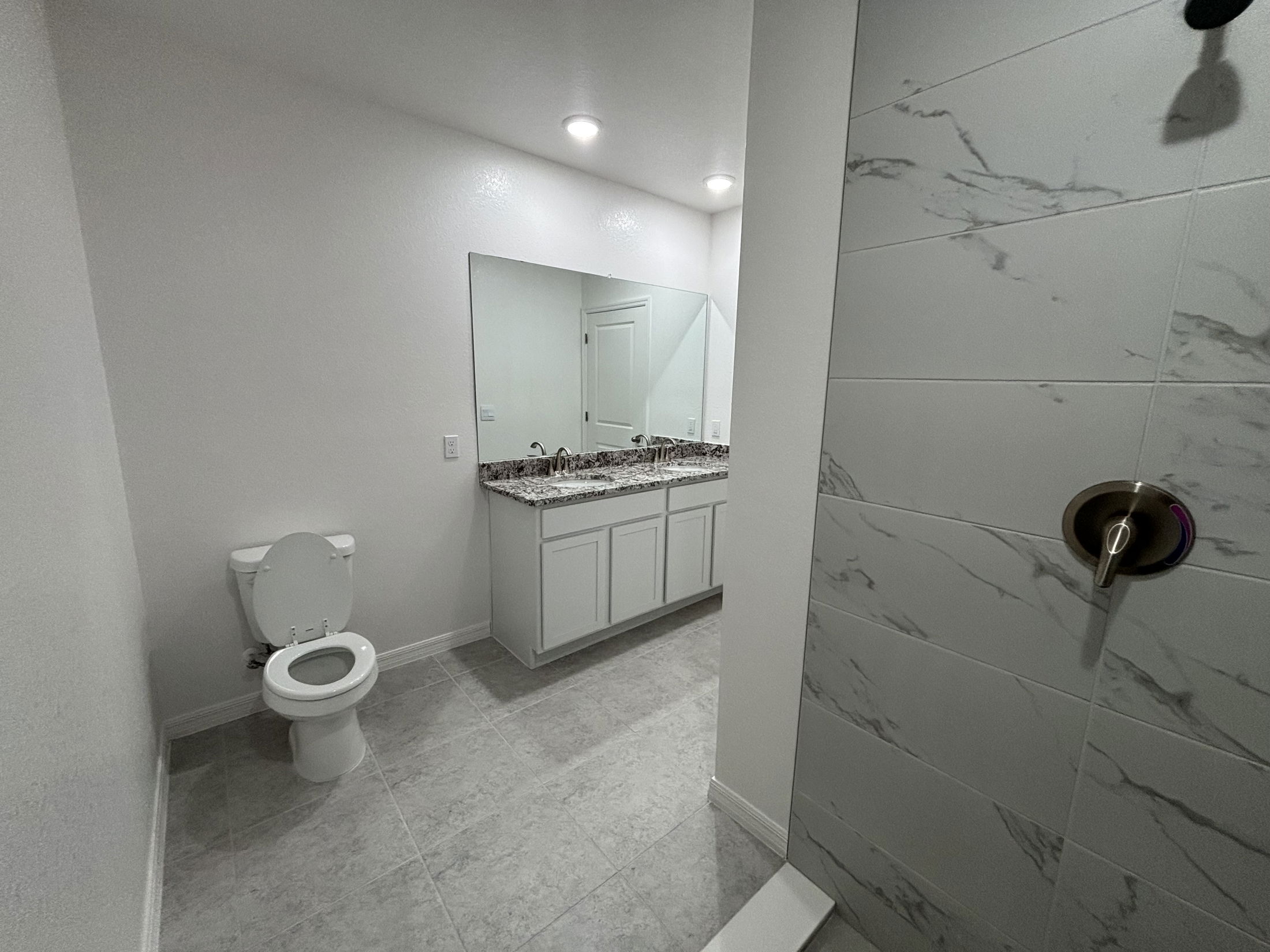 Modern bathroom interior featuring a white toilet, double sink vanity with granite countertop, large mirror, and a walk-in shower with marble tiles.