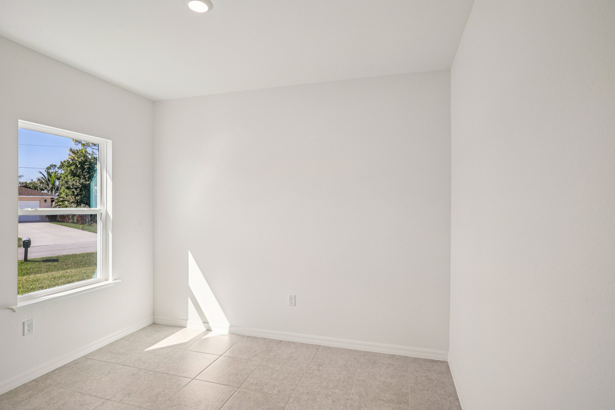 A bright, empty room with white walls and tile flooring, featuring a window that allows natural light to flood in.
