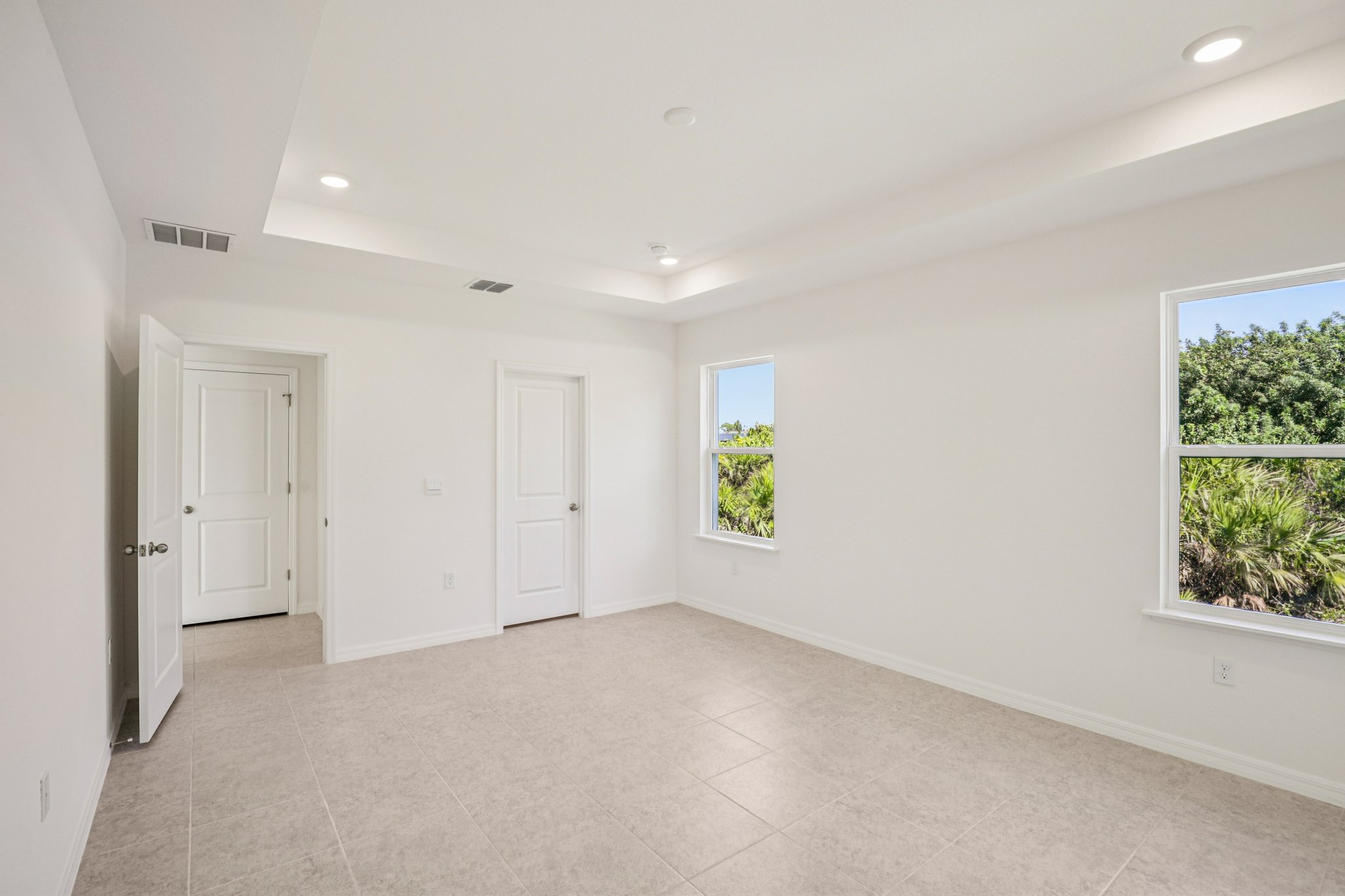 Bright and spacious empty room with white walls, tile flooring, and two windows overlooking greenery.