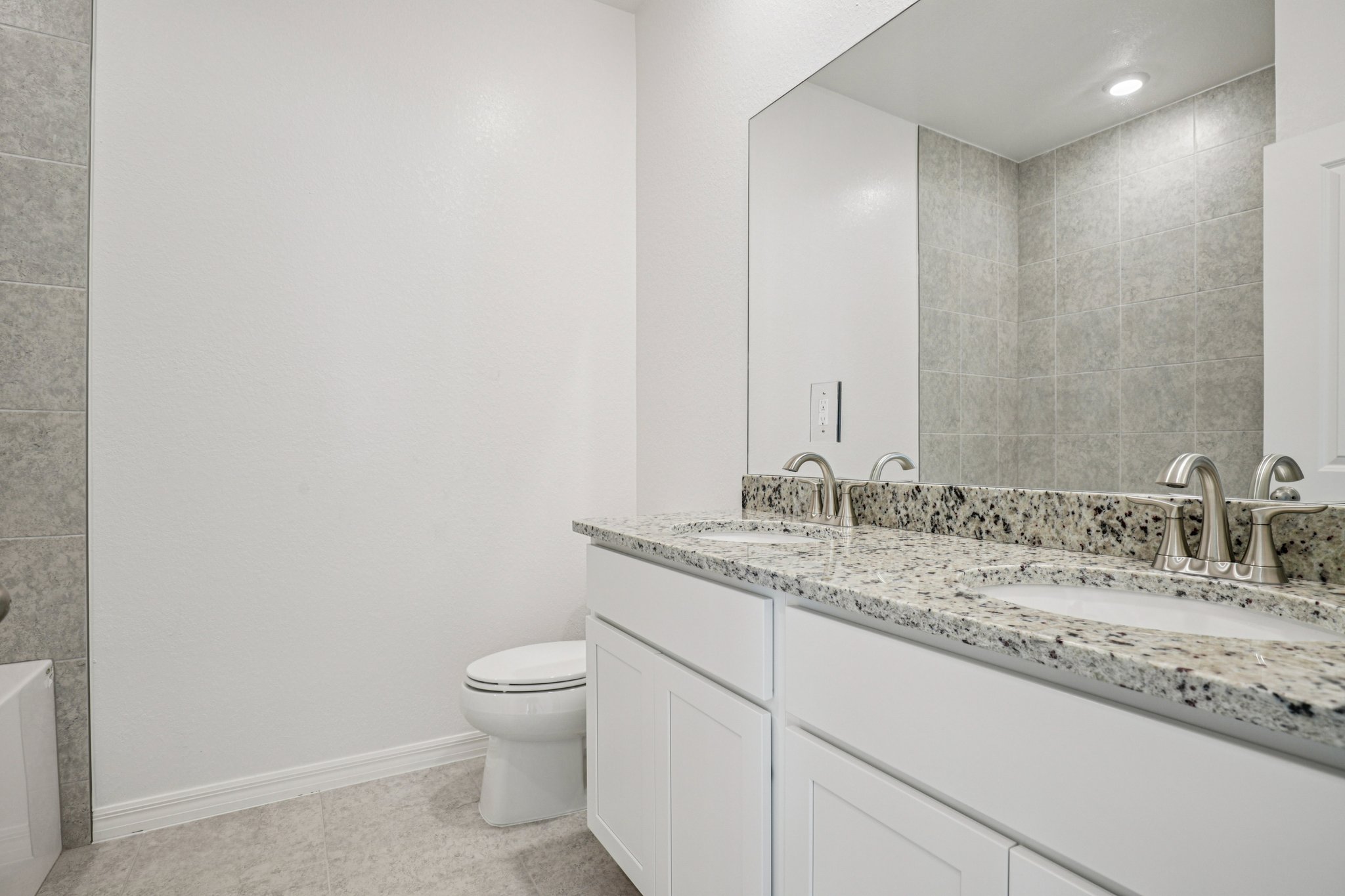 Modern bathroom featuring a double sink vanity with granite countertops, a spacious mirror, and gray tiled walls.