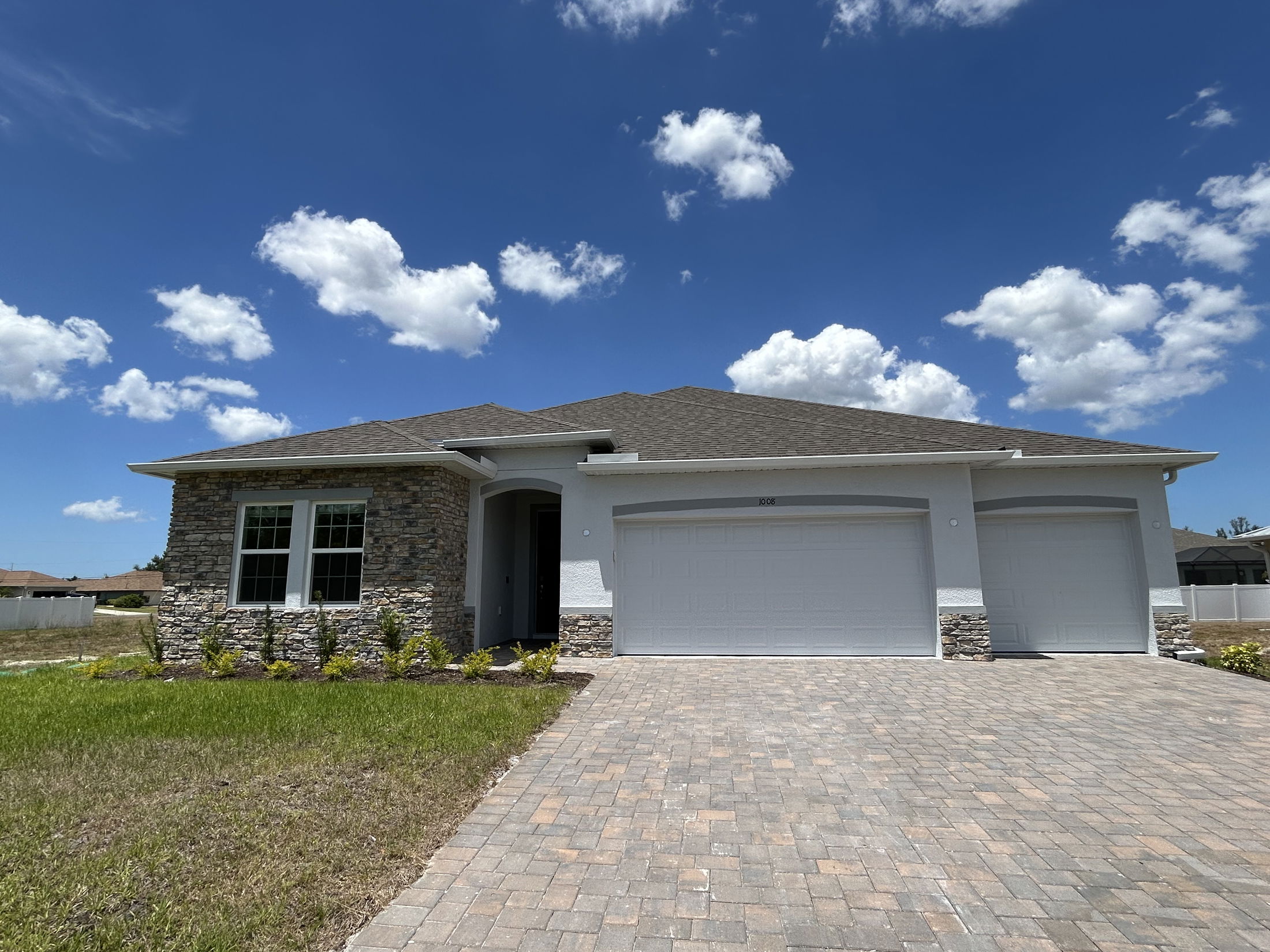 Modern single-story stone and stucco suburban house with a three-car garage and paved driveway under a clear blue sky.