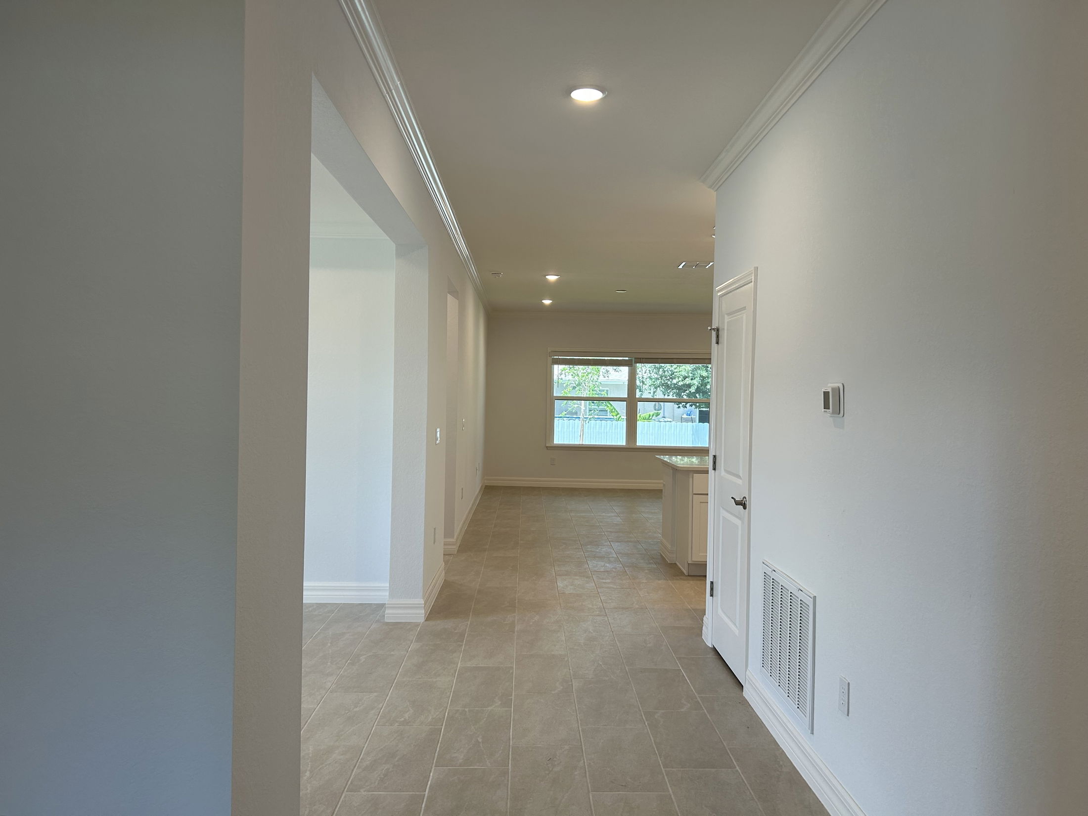 Spacious hallway in a modern home with tiled flooring leading to a brightly lit kitchen area.