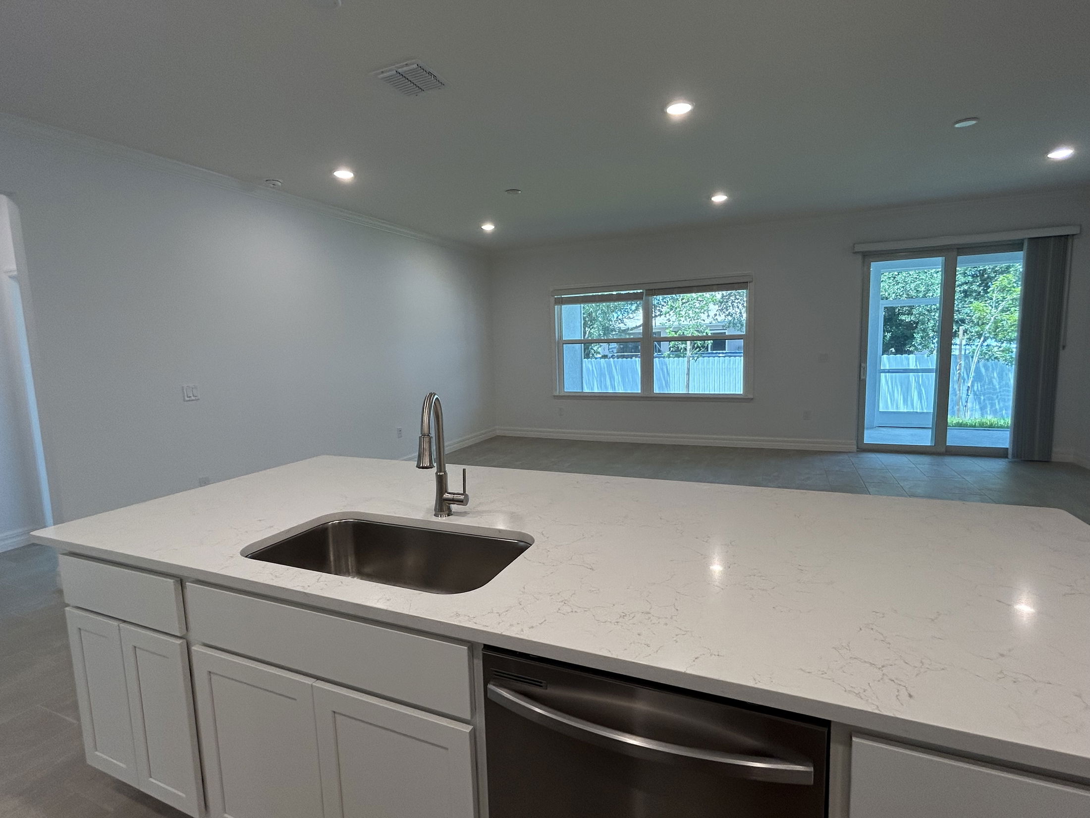 Spacious modern kitchen and living area with large windows, marble island, and stainless steel sink, featuring wooden floor and sliding glass doors leading to a backyard.