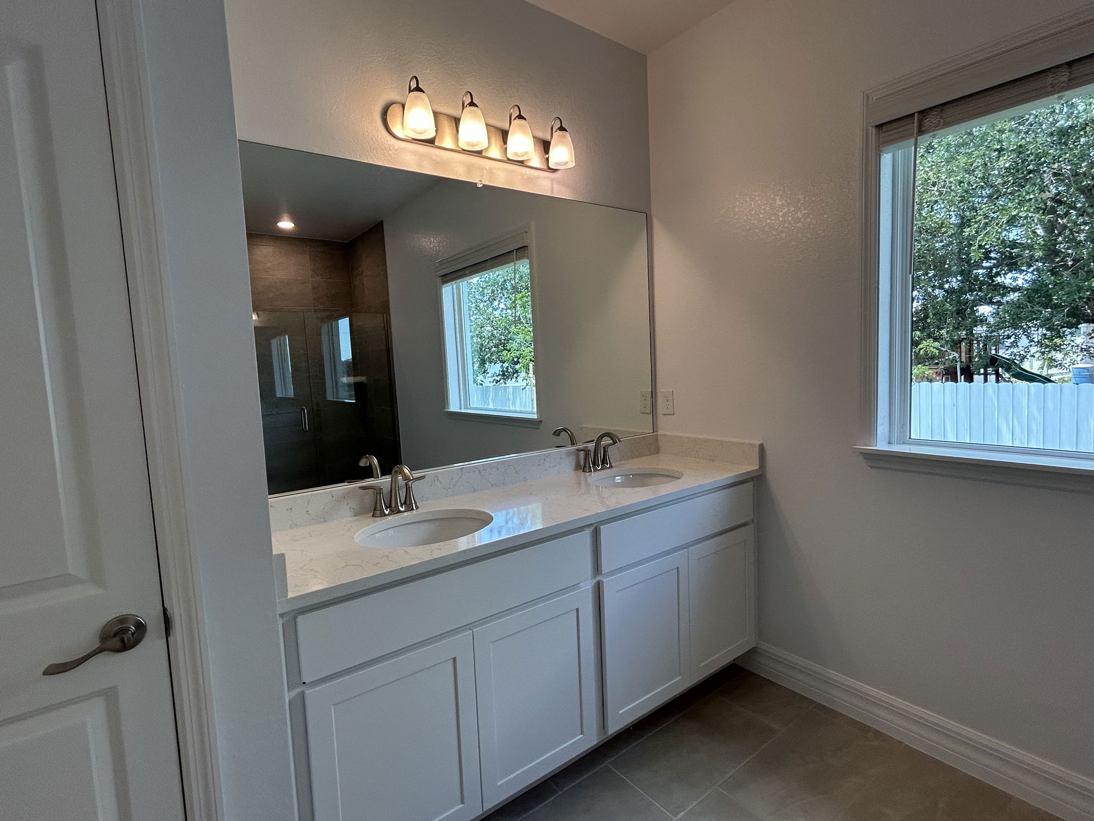 Modern bathroom with dual sinks, large mirror, contemporary lighting, and a window overlooking greenery.