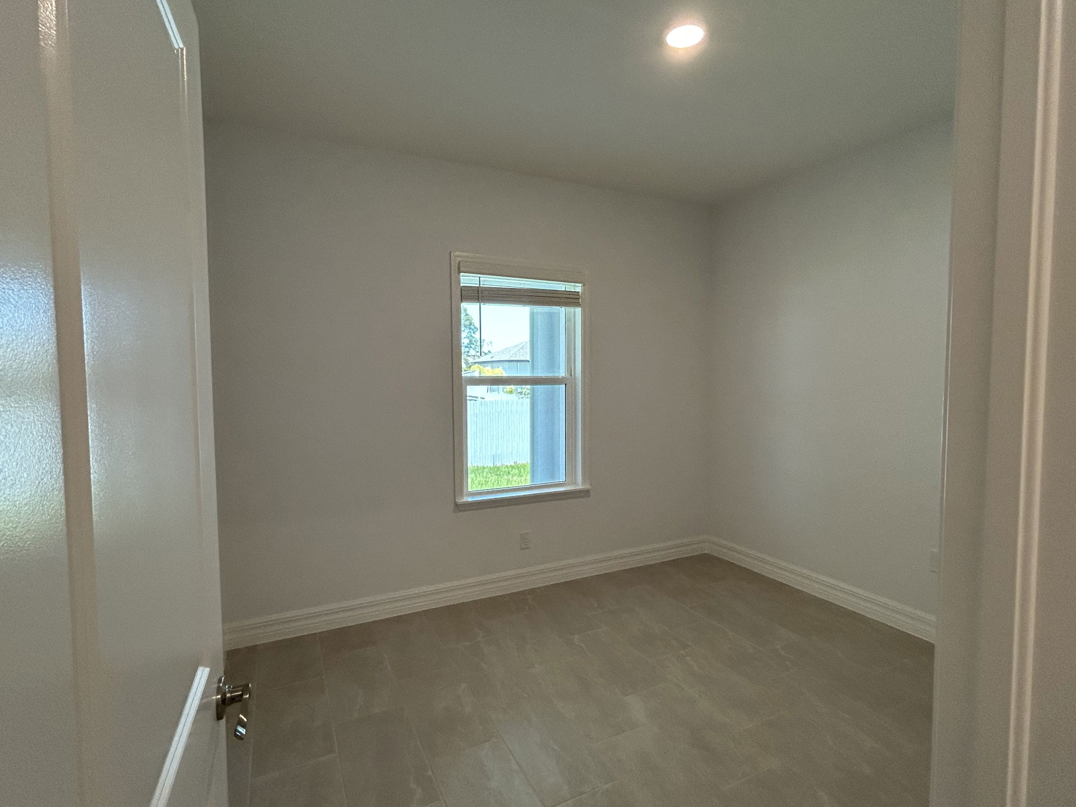 Empty room with beige tile flooring, white walls, and a single window letting in natural light.