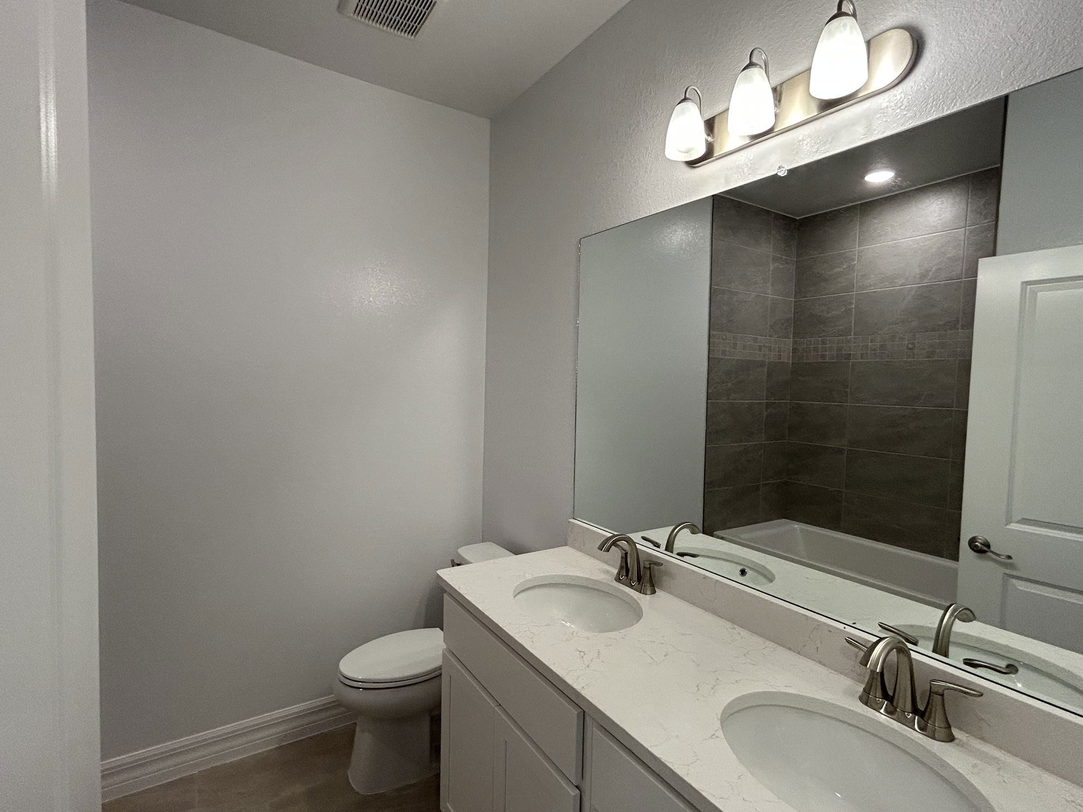 Modern bathroom with dual sinks, wall mirror, toilet, and a bathtub with gray tile surround under bright lighting.